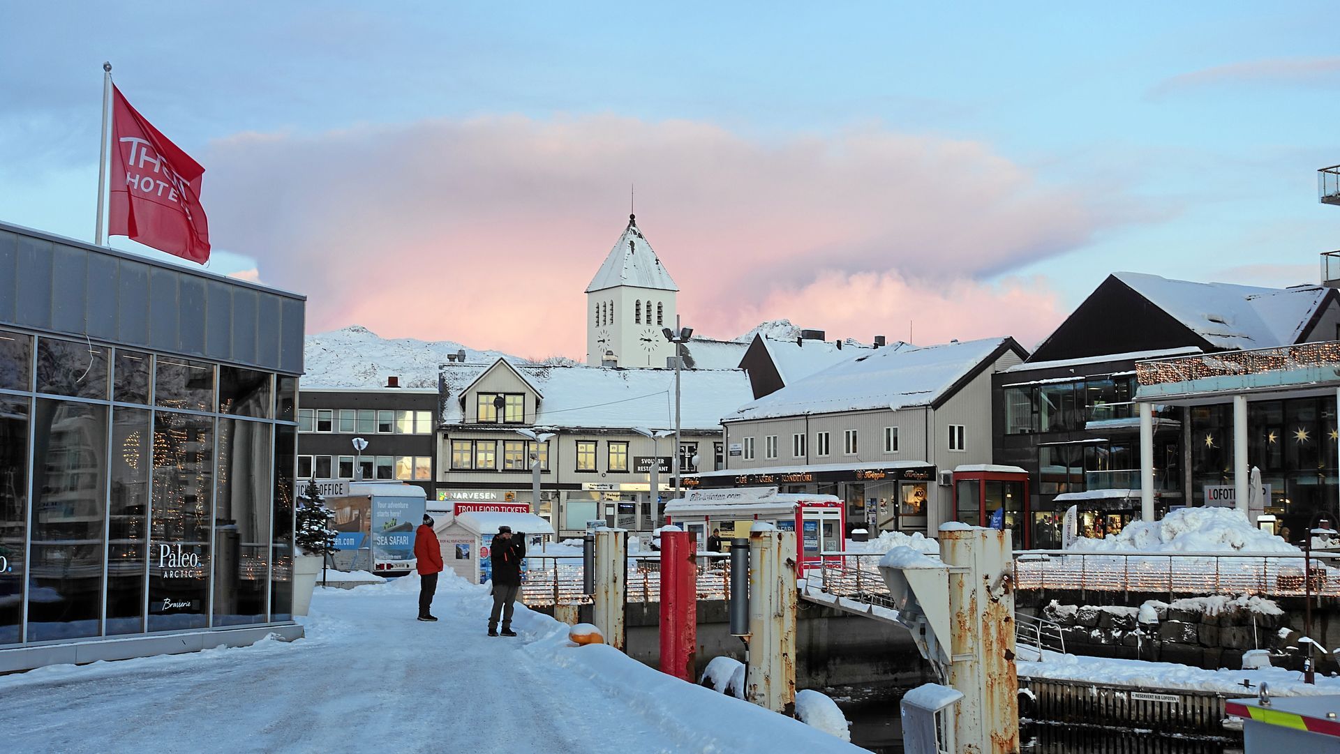 Verschneiter Marktplatz mit Gebäuden, Kirchturm und Menschen in der Nähe eines Piers in der Abenddämmerung. 