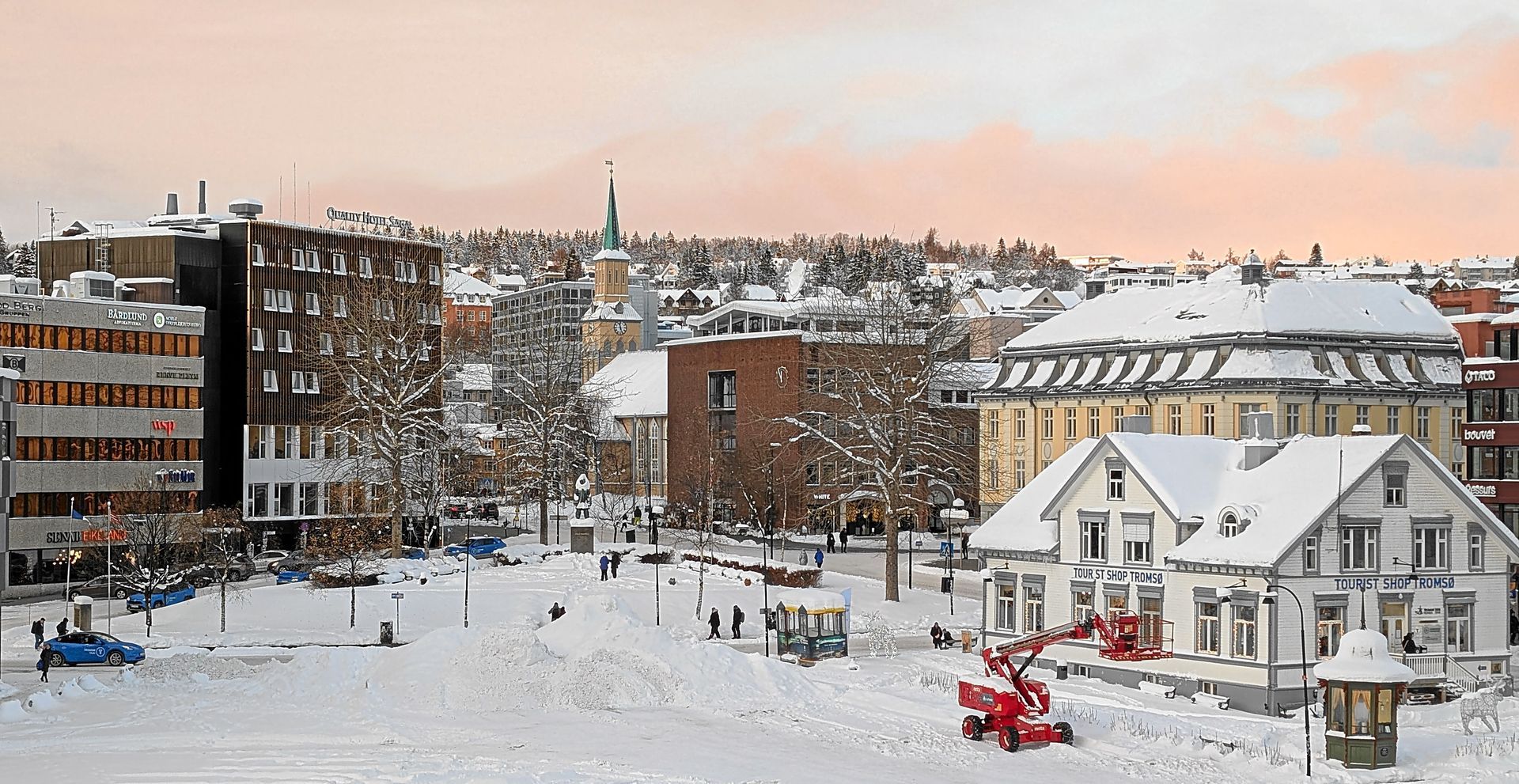 Schneebedecktes Stadtbild mit Gebäuden, einem Kirchturm und einer roten Schneefräse.