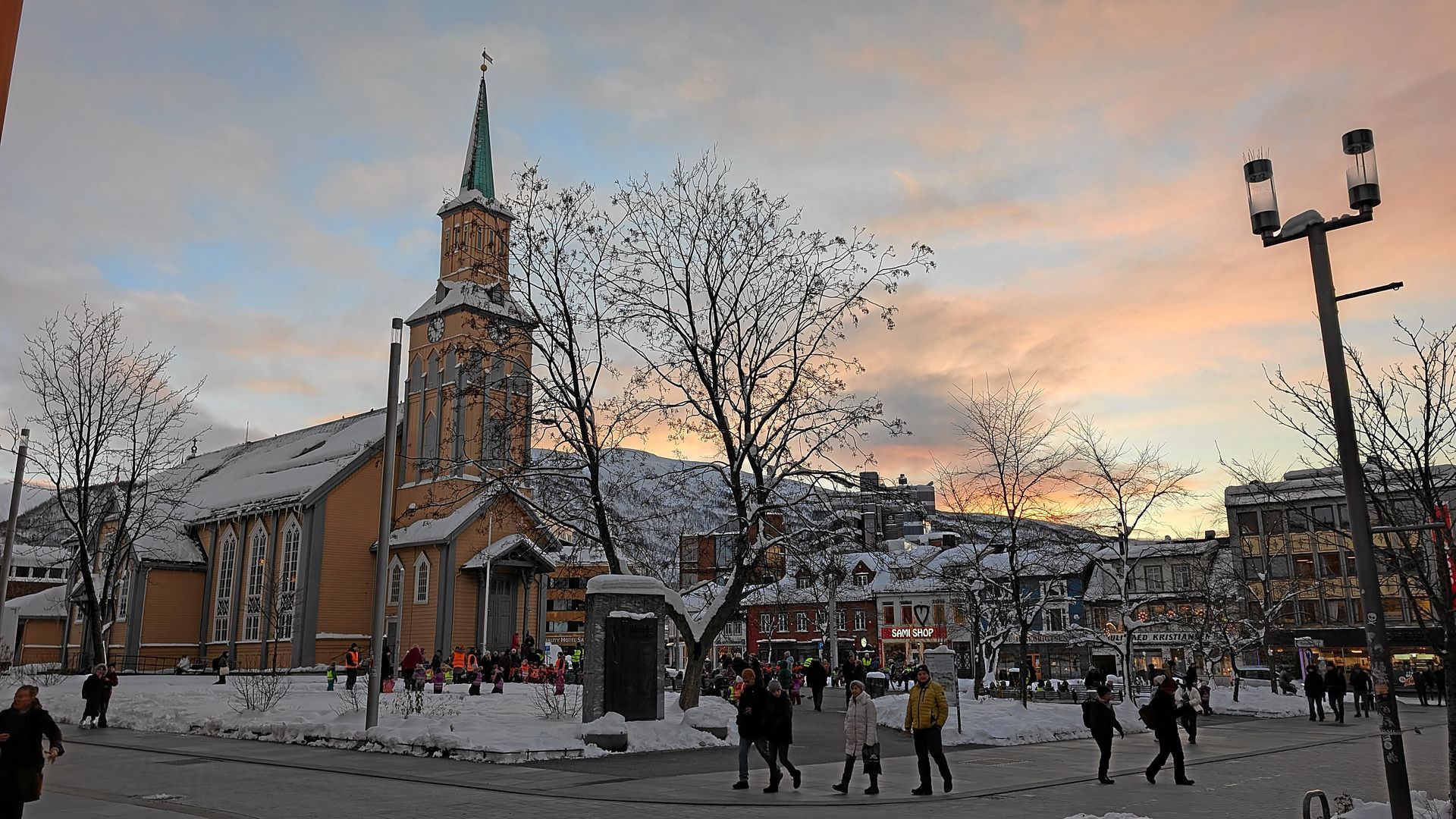 Kirche mit hohem Kirchturm auf einem schneebedeckten Platz bei Sonnenuntergang, Menschen gehen vorbei.