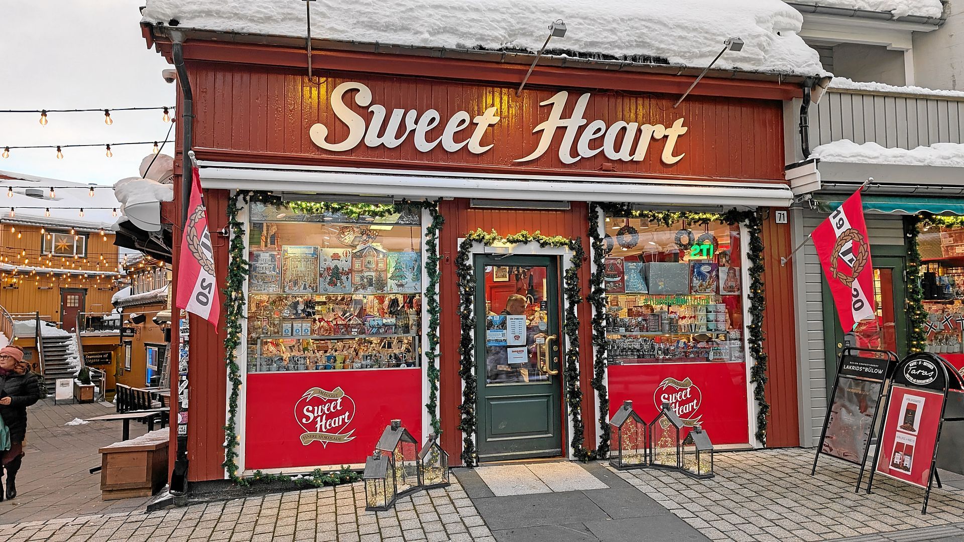 Außenansicht des Süßwarenladens „Sweet Heart“, rot-weiße Fassade, Schnee auf dem Dach, festlich geschmückt.