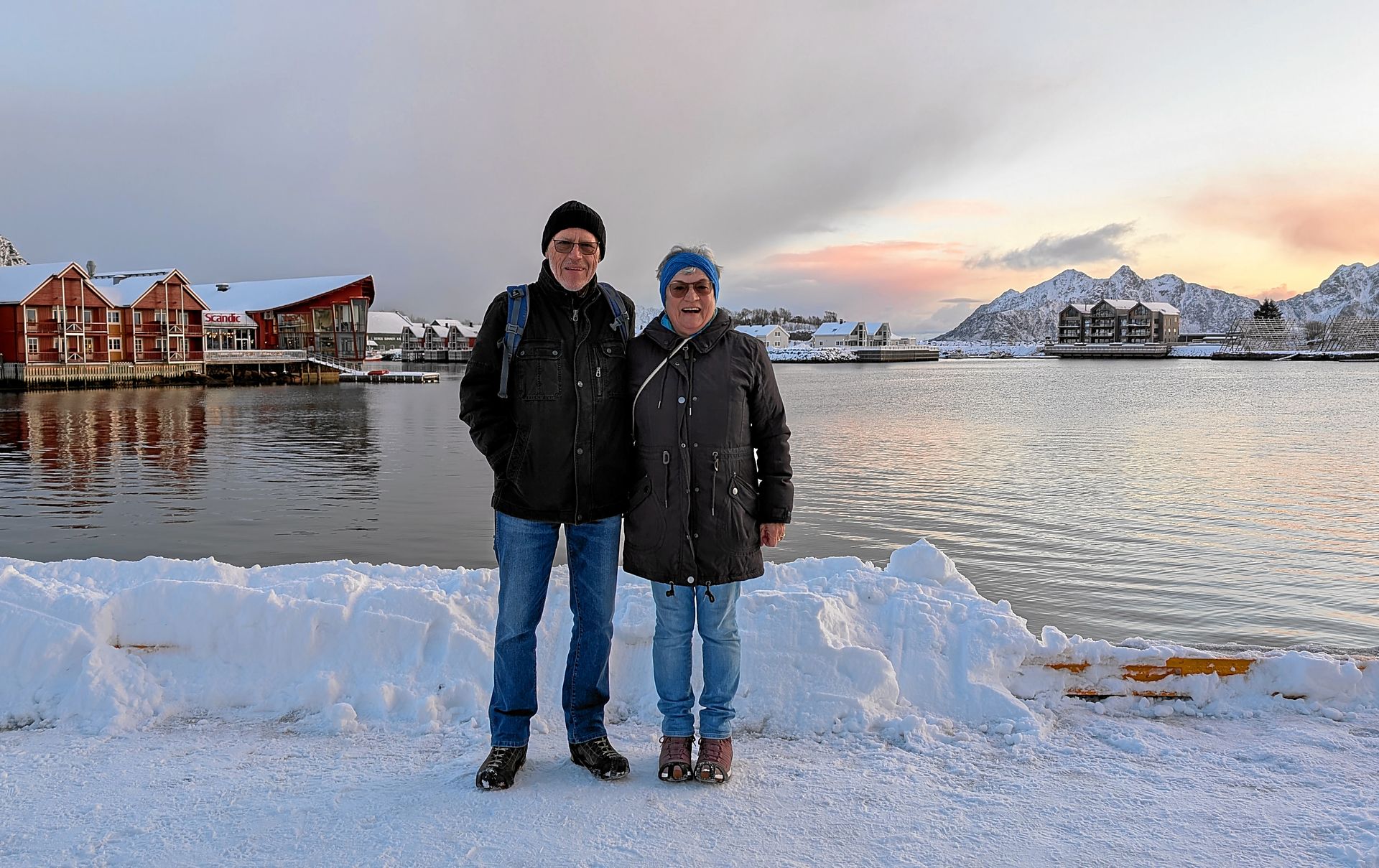 Zwei Personen stehen im Schnee, am Wasser, im Hintergrund Gebäude und Berge.