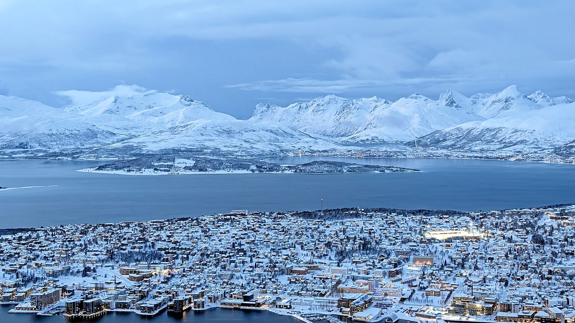 Eine schneebedeckte Stadt und Berge grenzen unter einem bewölkten Himmel an ein Gewässer.