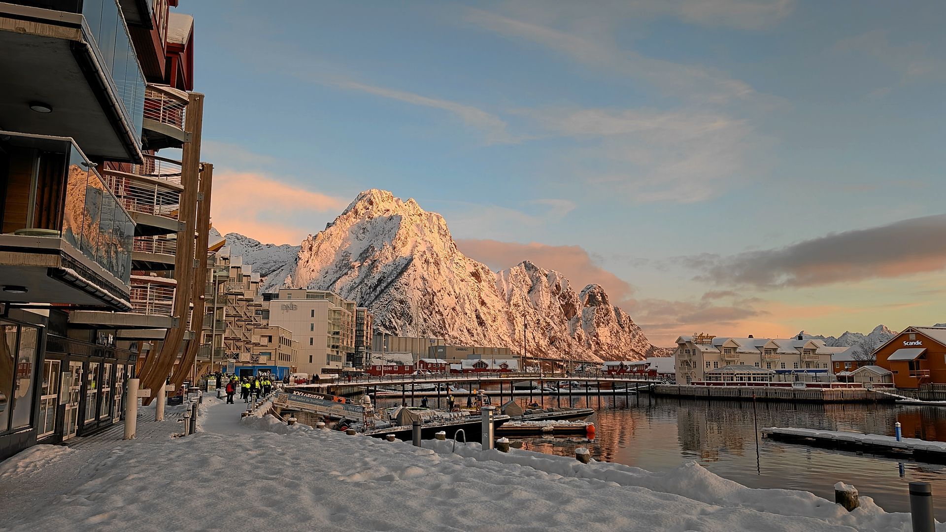 Verschneite Hafenszene mit Bergkulisse, Gebäuden und Booten im Sonnenuntergang.