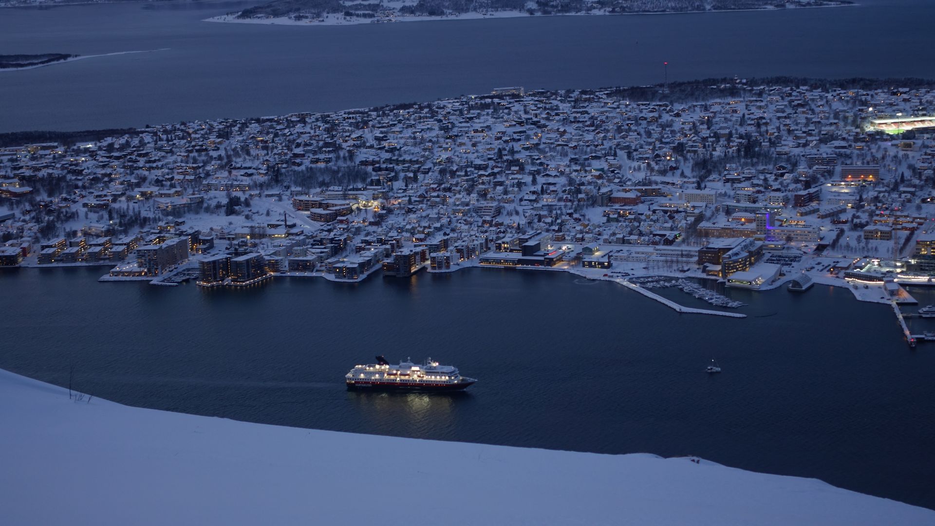 Ein Kreuzfahrtschiff fährt in der Abenddämmerung durch eine dunkelblaue Bucht auf eine schneebedeckte Stadt zu.