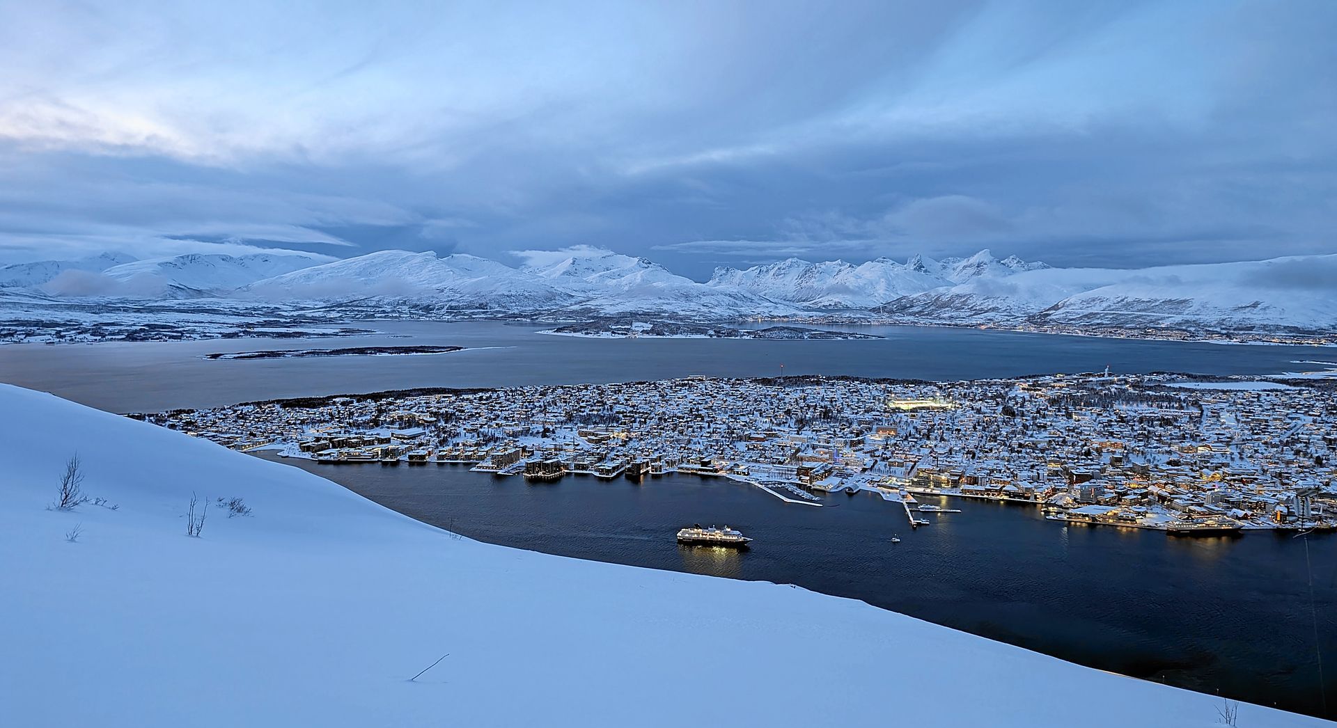 Eine schneebedeckte Stadt am Meer unter bewölktem Himmel mit schneebedeckten Bergen in der Ferne.