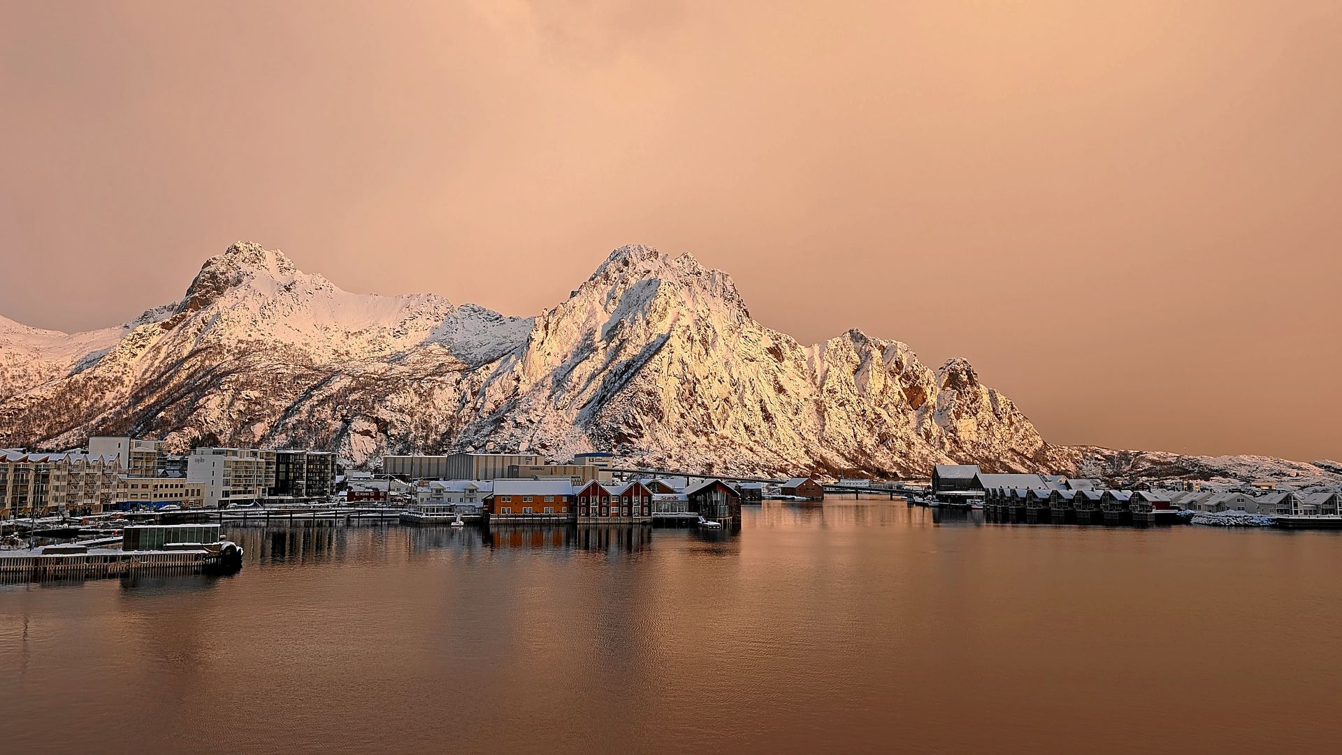Schneebedeckte Berge über einer Hafenstadt; lachsfarbener Himmel und Wasser, die die untergehende Sonne spiegeln.