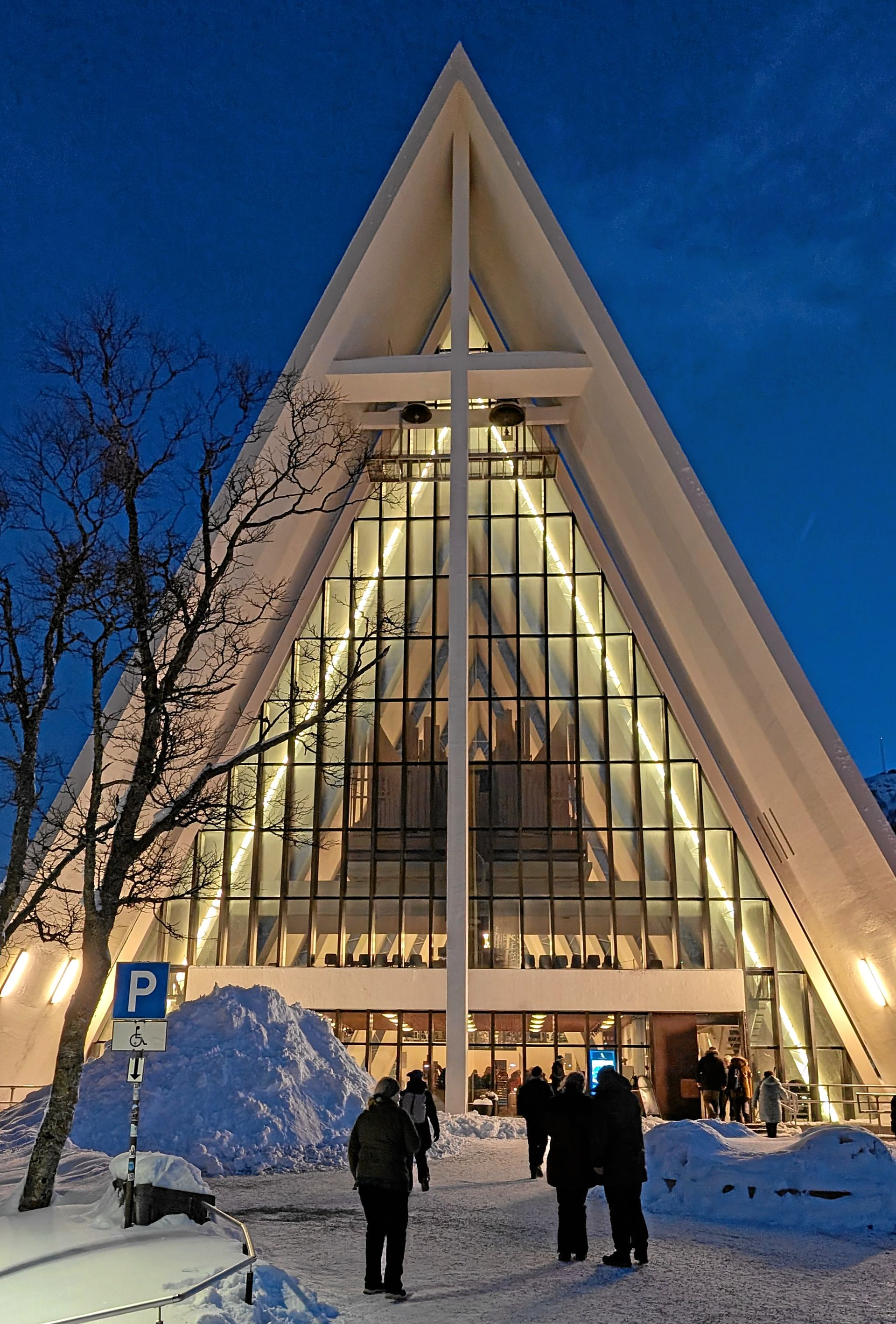 Eine Kathedrale mit dreieckigem Dach, Kreuz und Glasfassade, im Abendrot beleuchtet. Menschen sind im Vordergrund zu sehen.