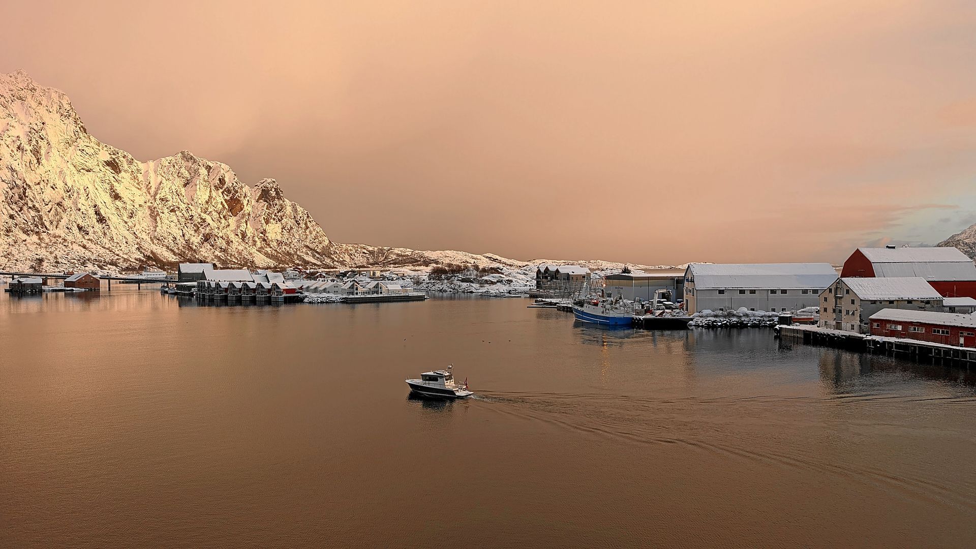 Verschneite Hafenstadt mit Bergen im Hintergrund unter dämmerigem Himmel, mit Booten und Gebäuden.