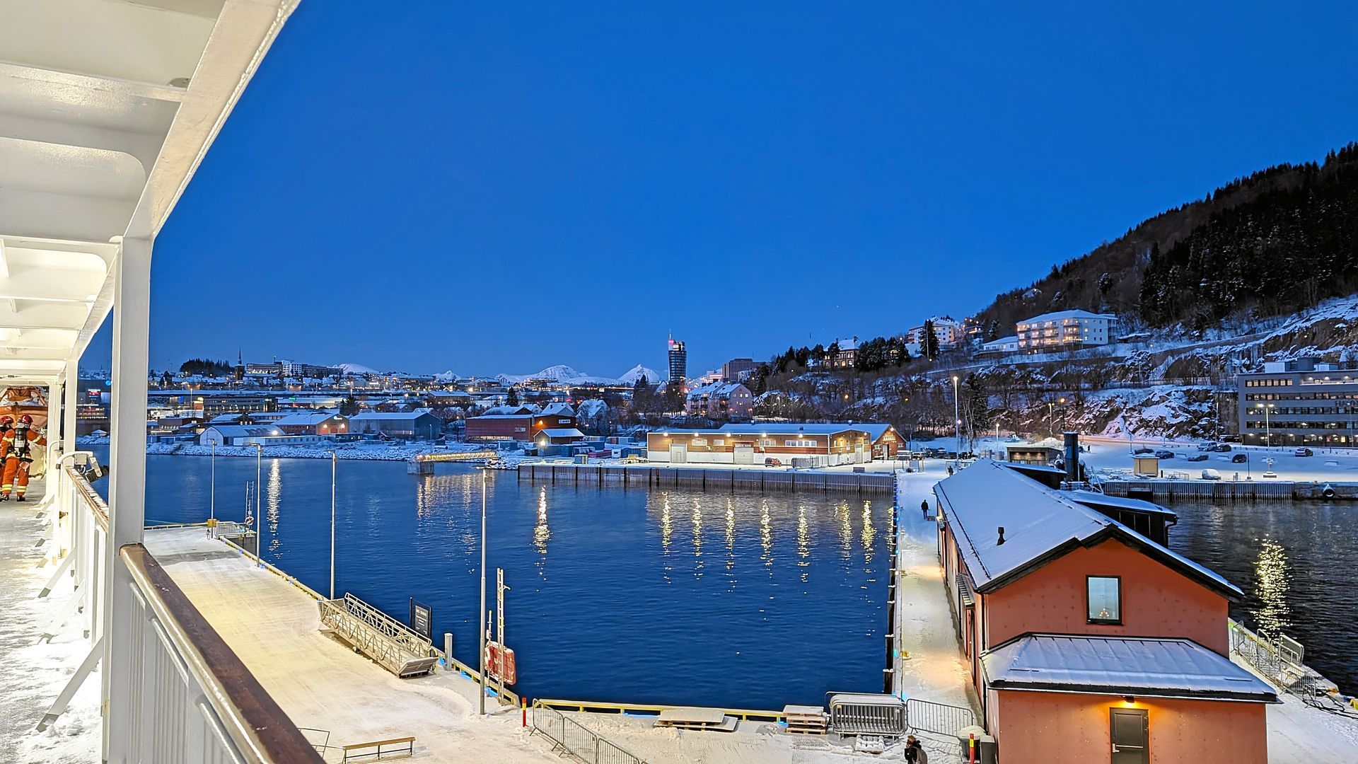 Eine verschneite Hafenszene in der Abenddämmerung. Gebäude und Lichter spiegeln sich im dunklen Wasser. Im Vordergrund ist das Deck eines Schiffes zu sehen.
