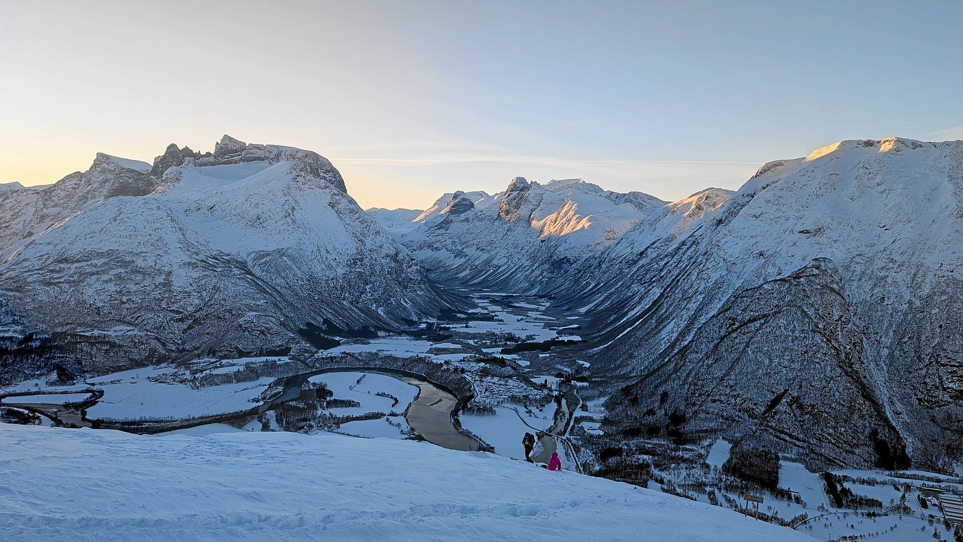 Bei Sonnenuntergang umgeben schneebedeckte Berge ein Tal mit einer Stadt und einem Fluss.