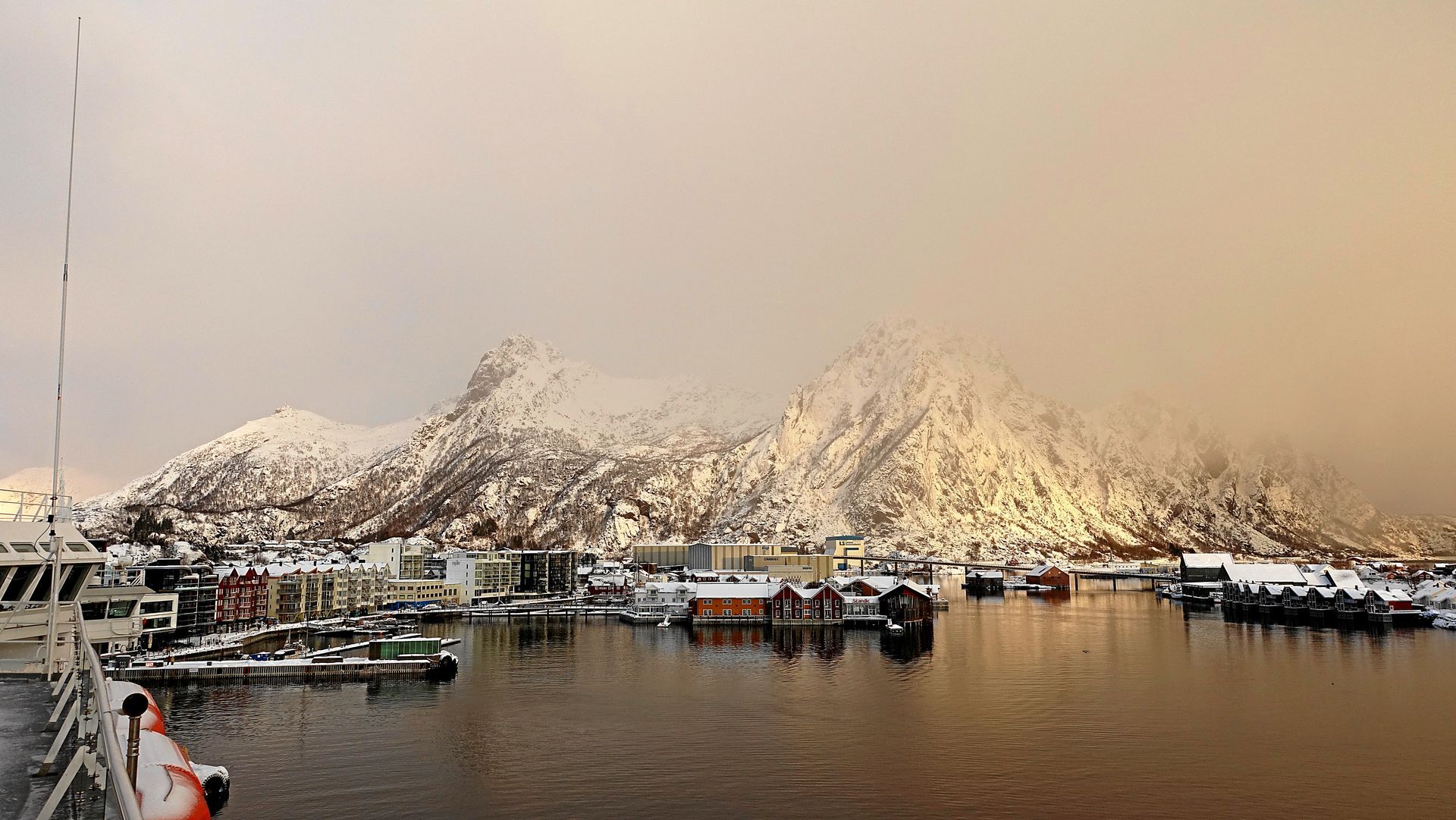 Verschneite Hafenszene in Norwegen mit Bergen, Gebäuden und Booten unter bedecktem Himmel.