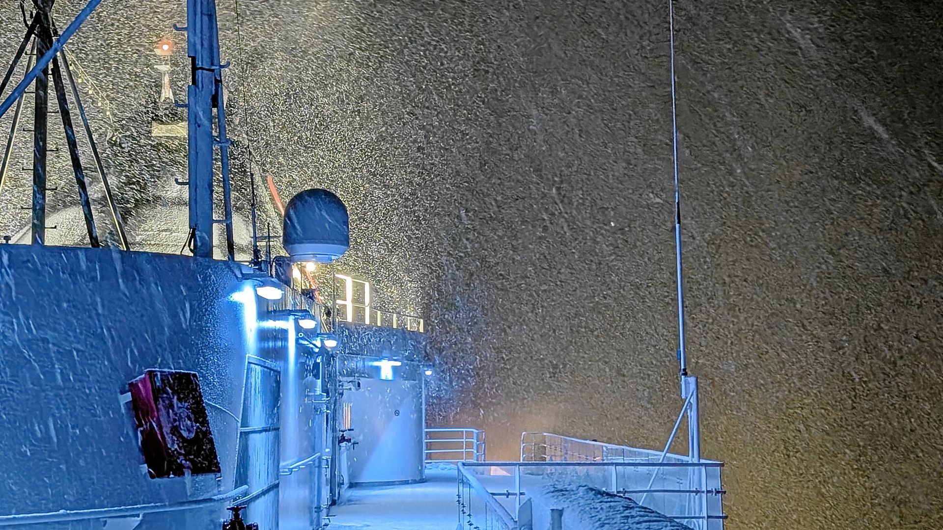 Schiffsdeck im Schneesturm, von Lichtern erhellt. Dunkler Himmel mit wirbelnden Schneeflocken.