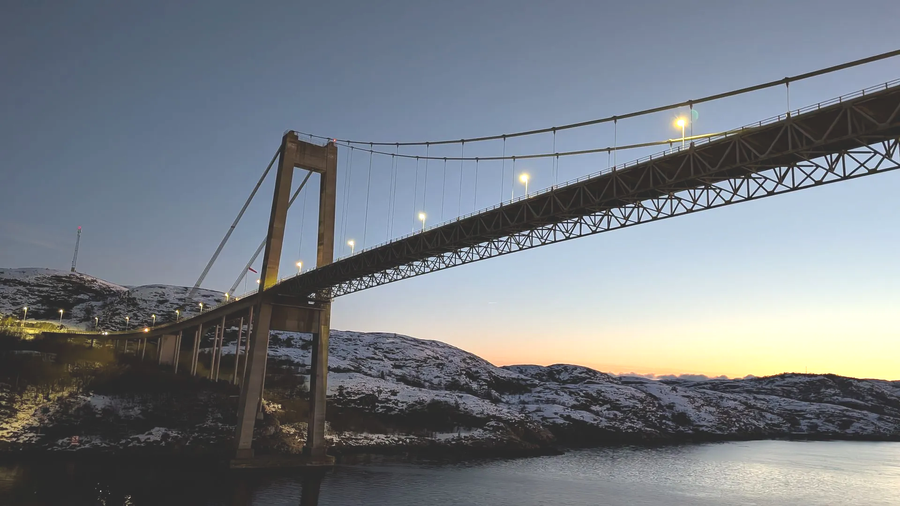Hängebrücke über Wasser in der Abenddämmerung, im Hintergrund schneebedeckte Hügel.