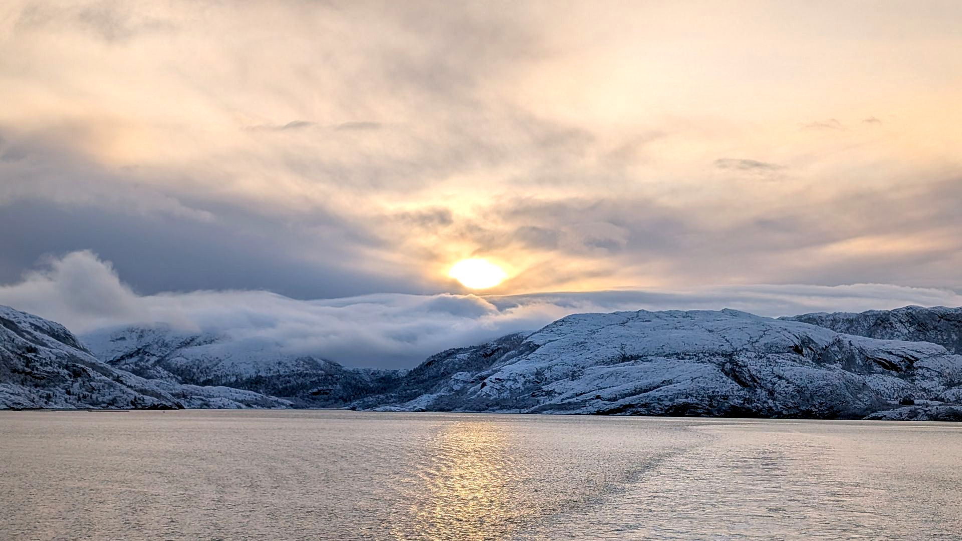 Verschneite Berge und Wasser unter einem bewölkten Himmel, durch dessen Wolken die Sonne hindurchlugt.