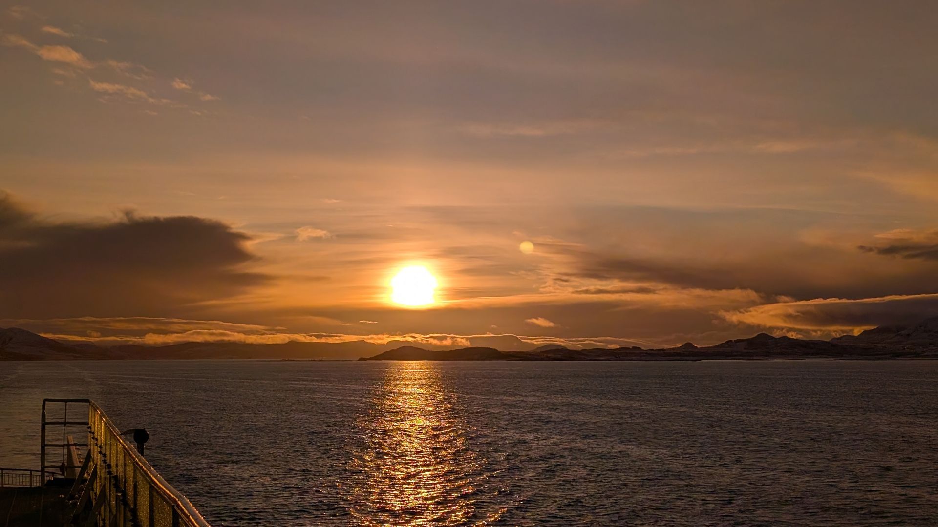 Sonnenuntergang über dem Meer; orange und goldene Farbtöne spiegeln sich im Wasser. Dunkle Wolken und Land in der Ferne.