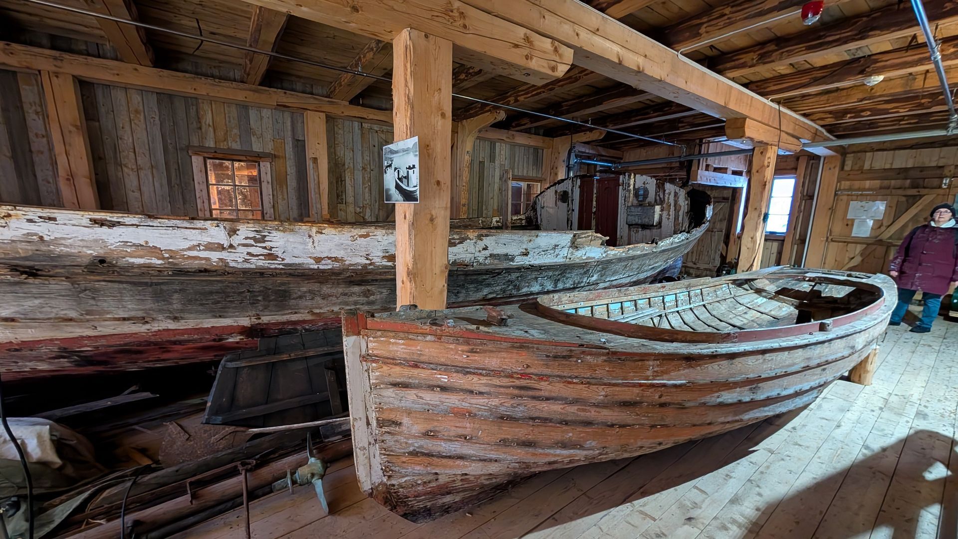Alte Holzboote sind in einem verwitterten Holzgebäude ausgestellt. Eine Person steht daneben.