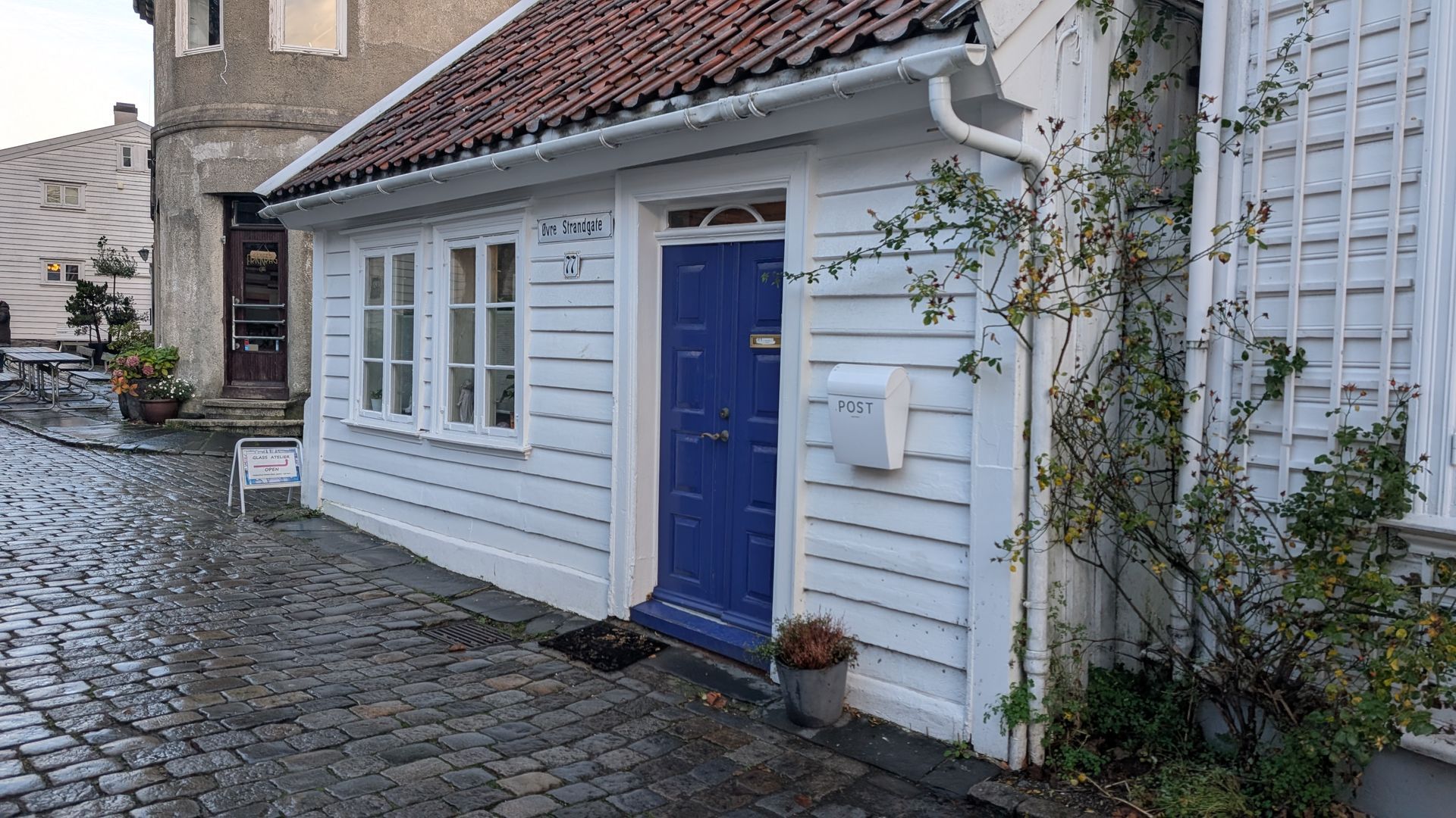 Weißes Holzhäuschen mit blauer Tür, rotem Ziegeldach und Kopfsteinpflasterstraße.