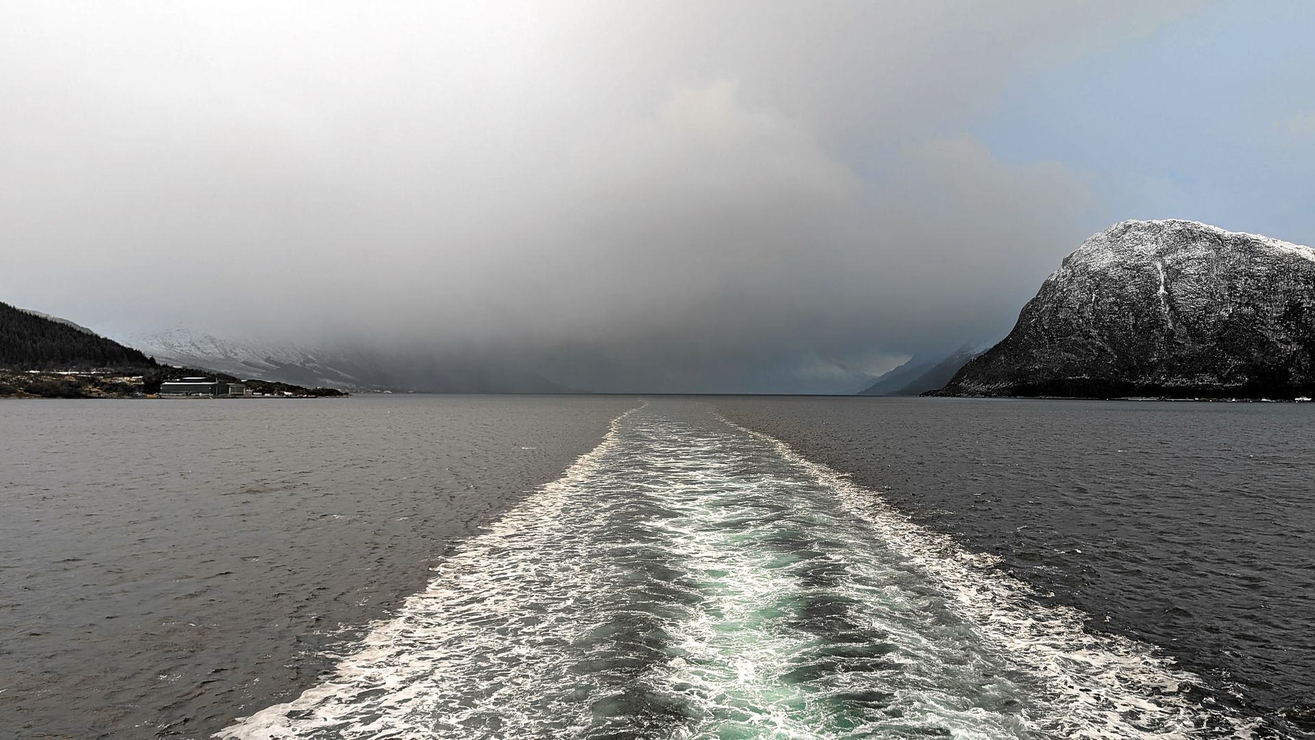 Kielwasser eines Bootes auf dunklem Wasser, Berge und ein bewölkter Himmel in der Ferne.