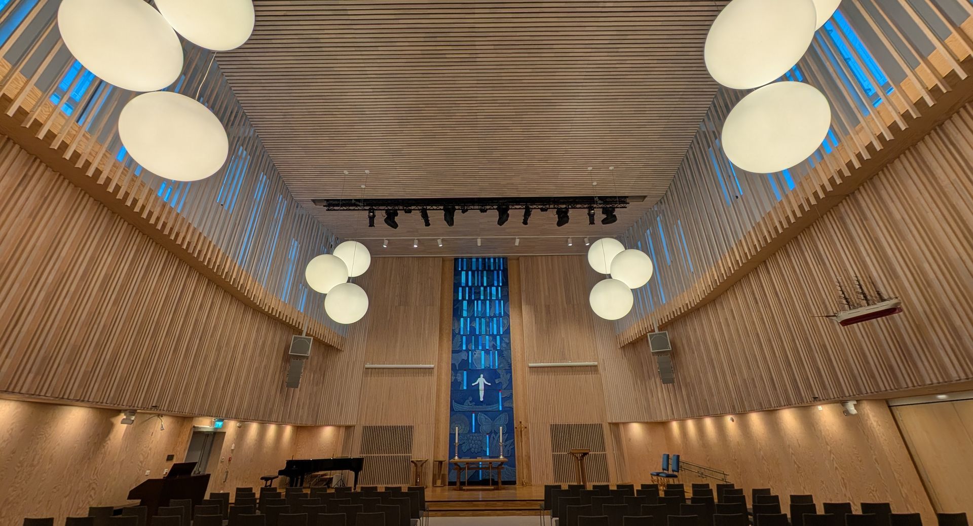 Innenansicht einer modernen Kirche mit Holzvertäfelung und großen Kugellampen. Über dem Altar ist ein Kreuz zu sehen.