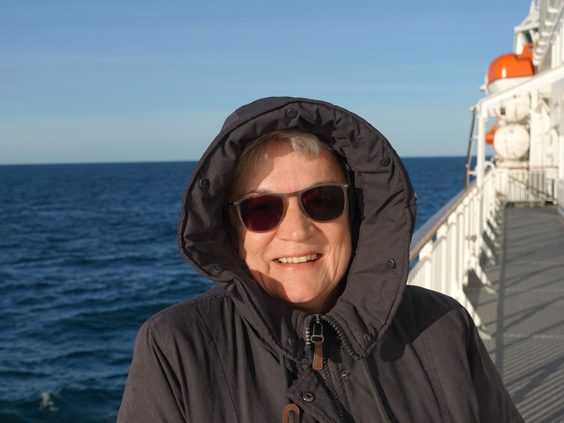 Eine Frau in Kapuzenjacke und Sonnenbrille lächelt auf einem Boot, im Hintergrund ist das Meer zu sehen.