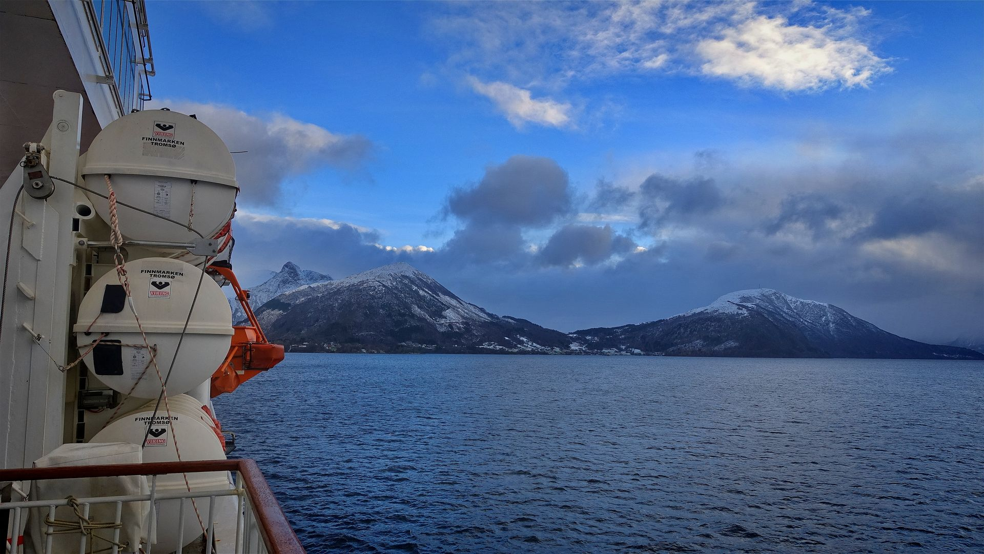 Bootsdeck mit Rettungsflößen, Blick auf schneebedeckte Berge und Meer unter bewölktem Himmel.