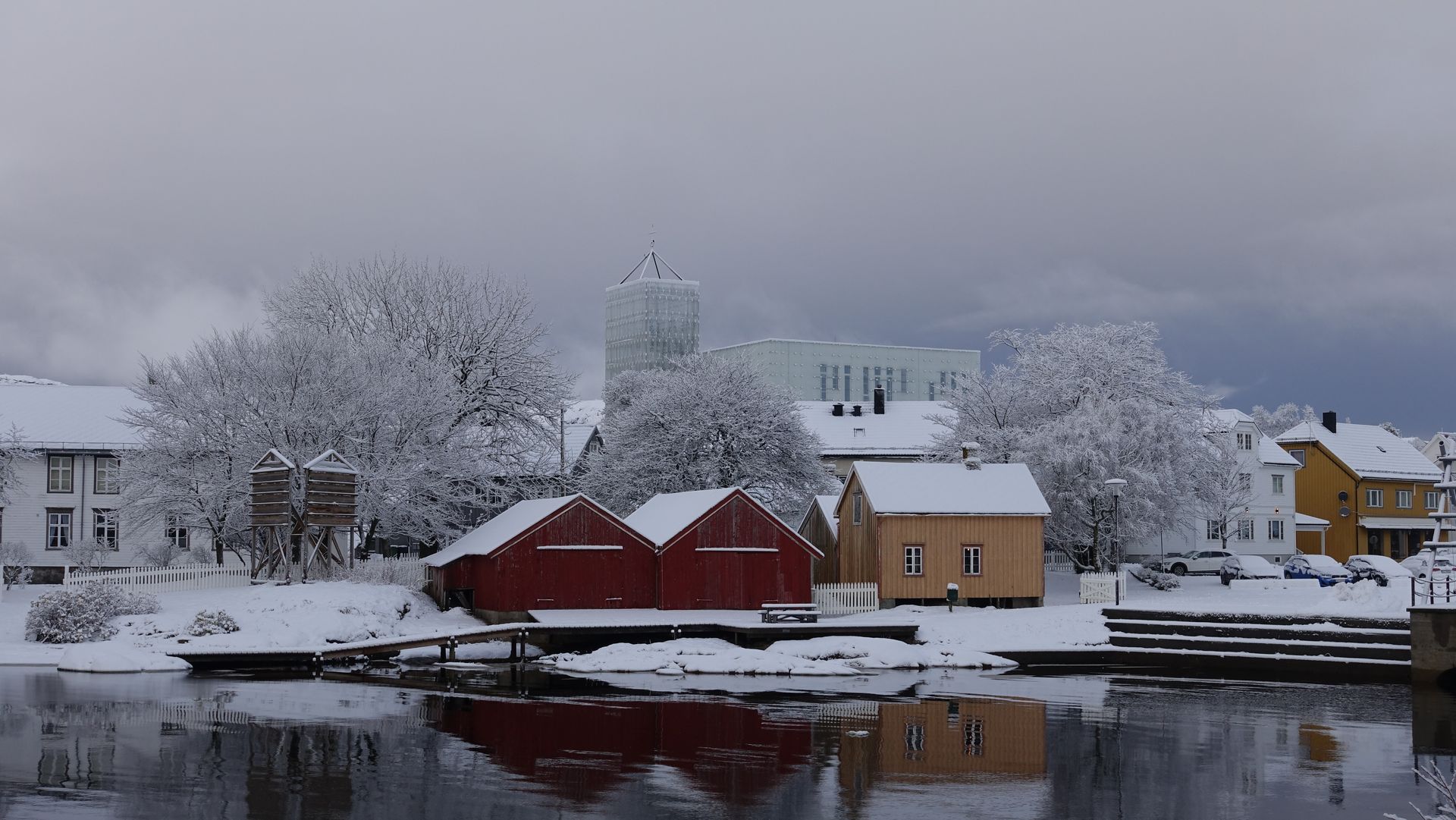 In einer winterlichen Szene säumen schneebedeckte Gebäude und Bäume ein Ufer. Rote Gebäude  spiegeln sich im Wasser.
