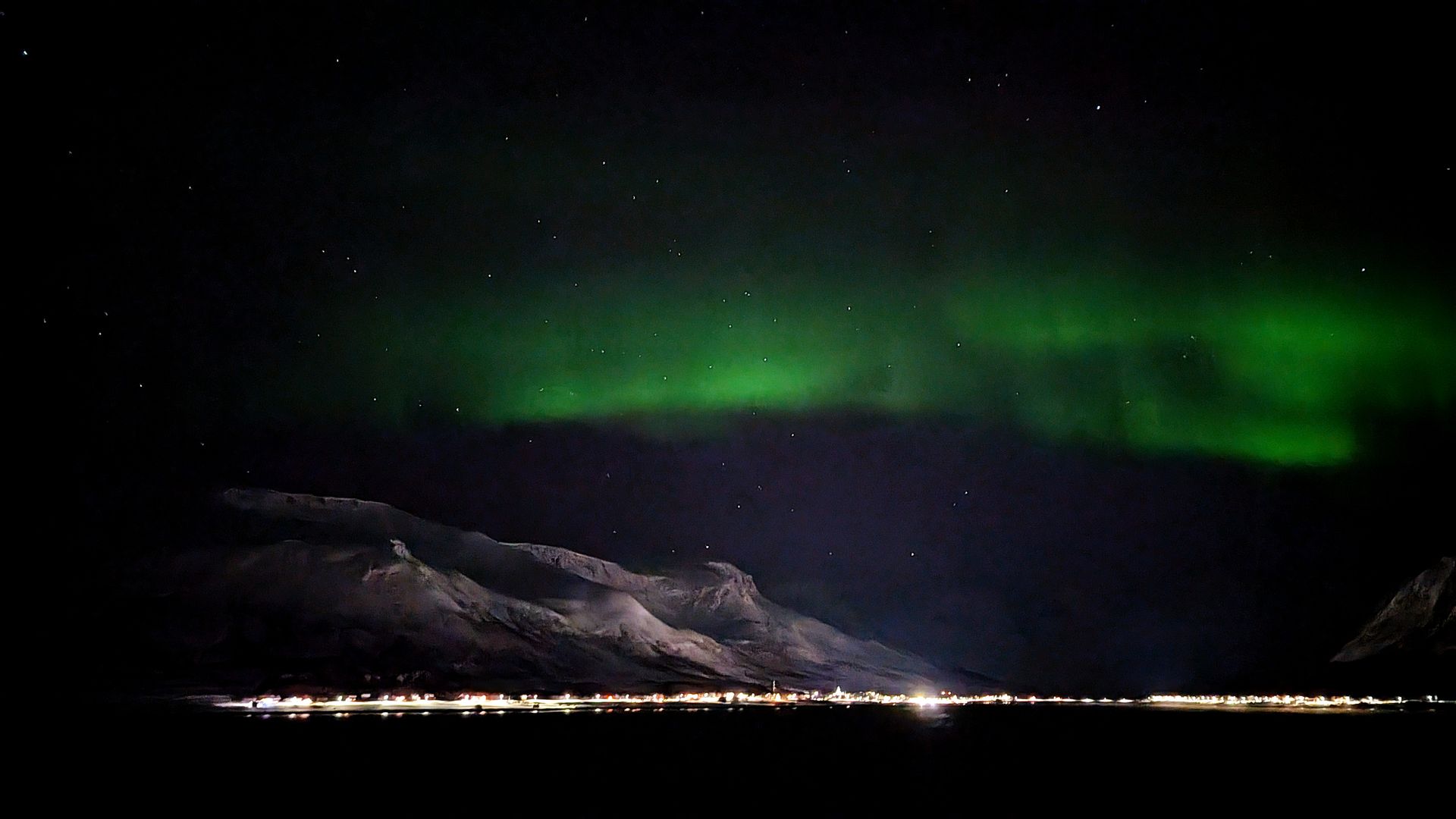 Grüne Nordlichter zeigen sich nachts über einer Bergkette und einer fernen, beleuchteten Küstenlinie.