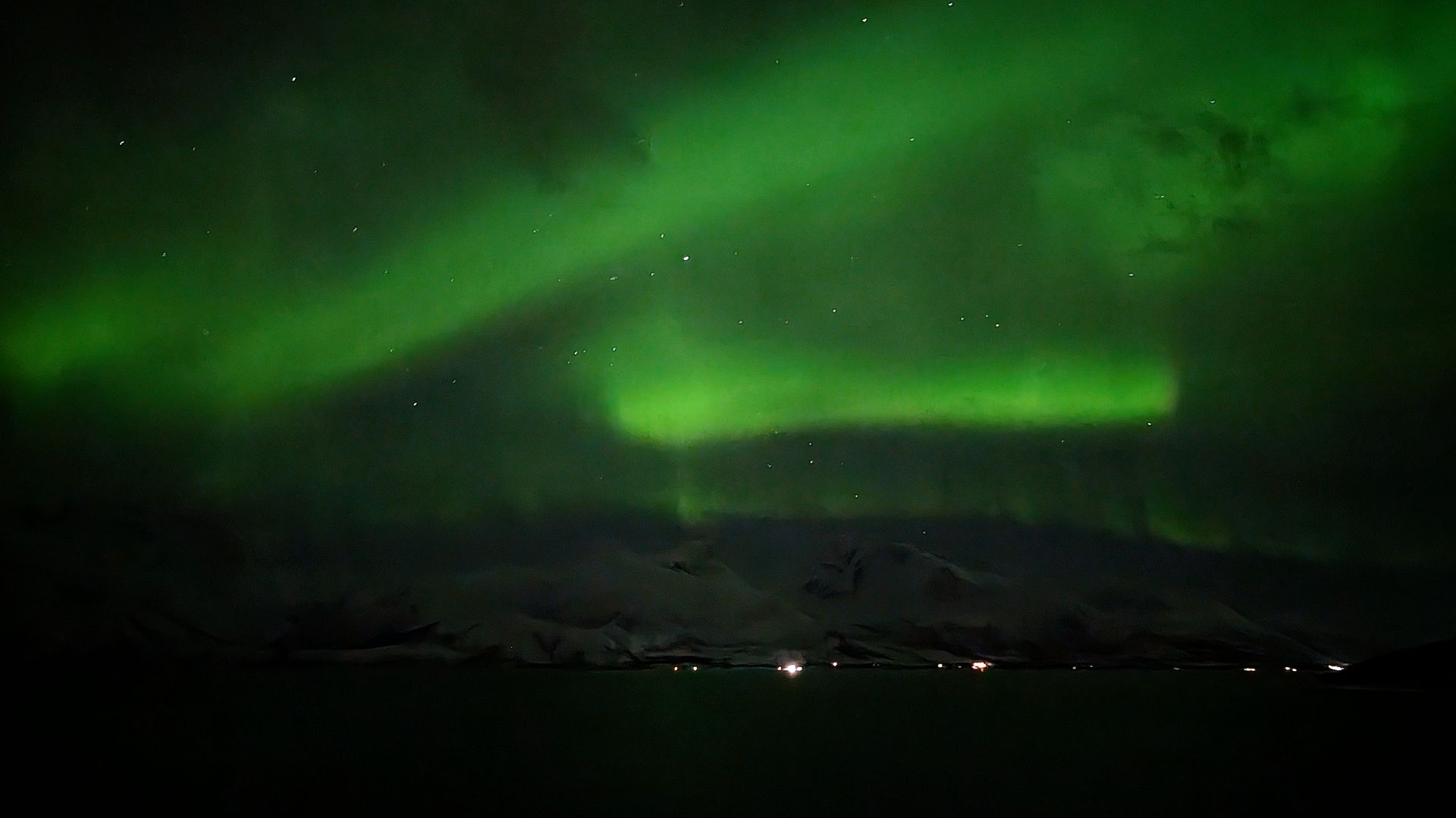 Grüne Nordlichter leuchten über dunklen Bergen und Gewässern.