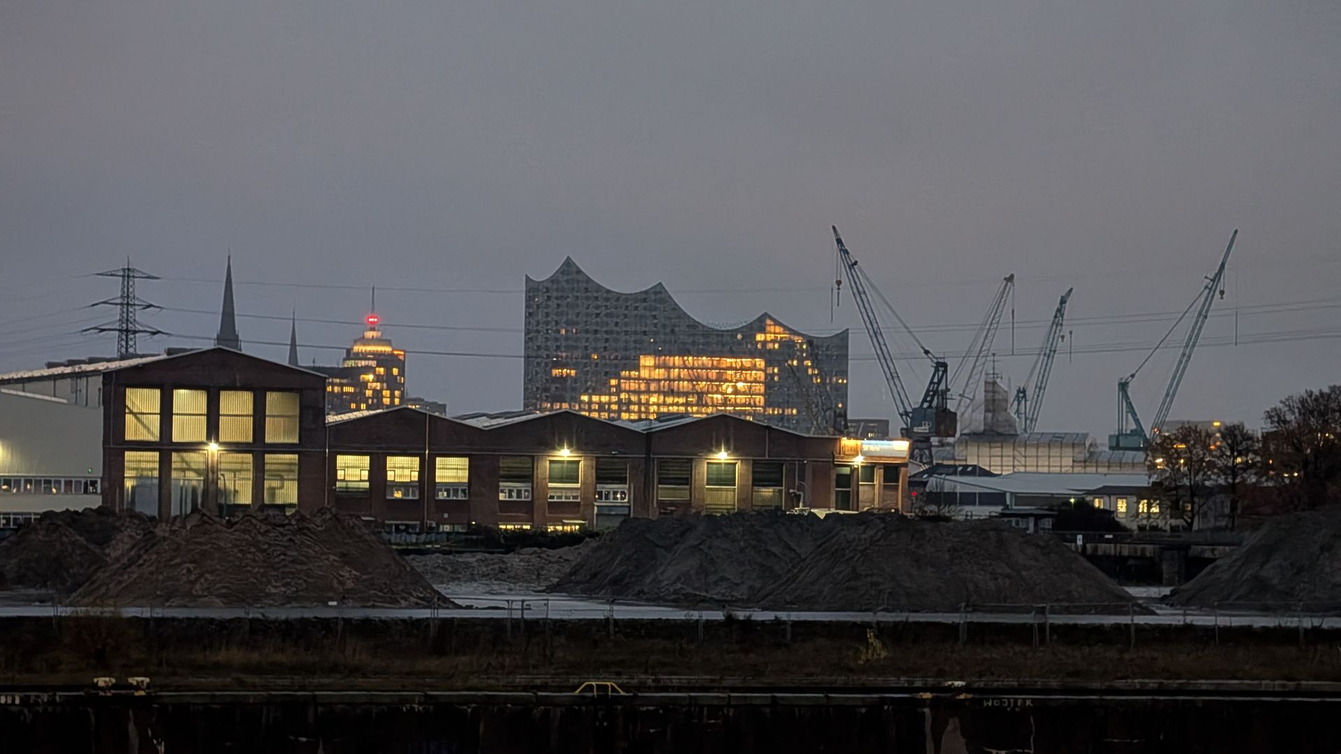 Abendliches Stadtbild mit beleuchteten Gebäuden, Kränen und der Elbphilharmonie.