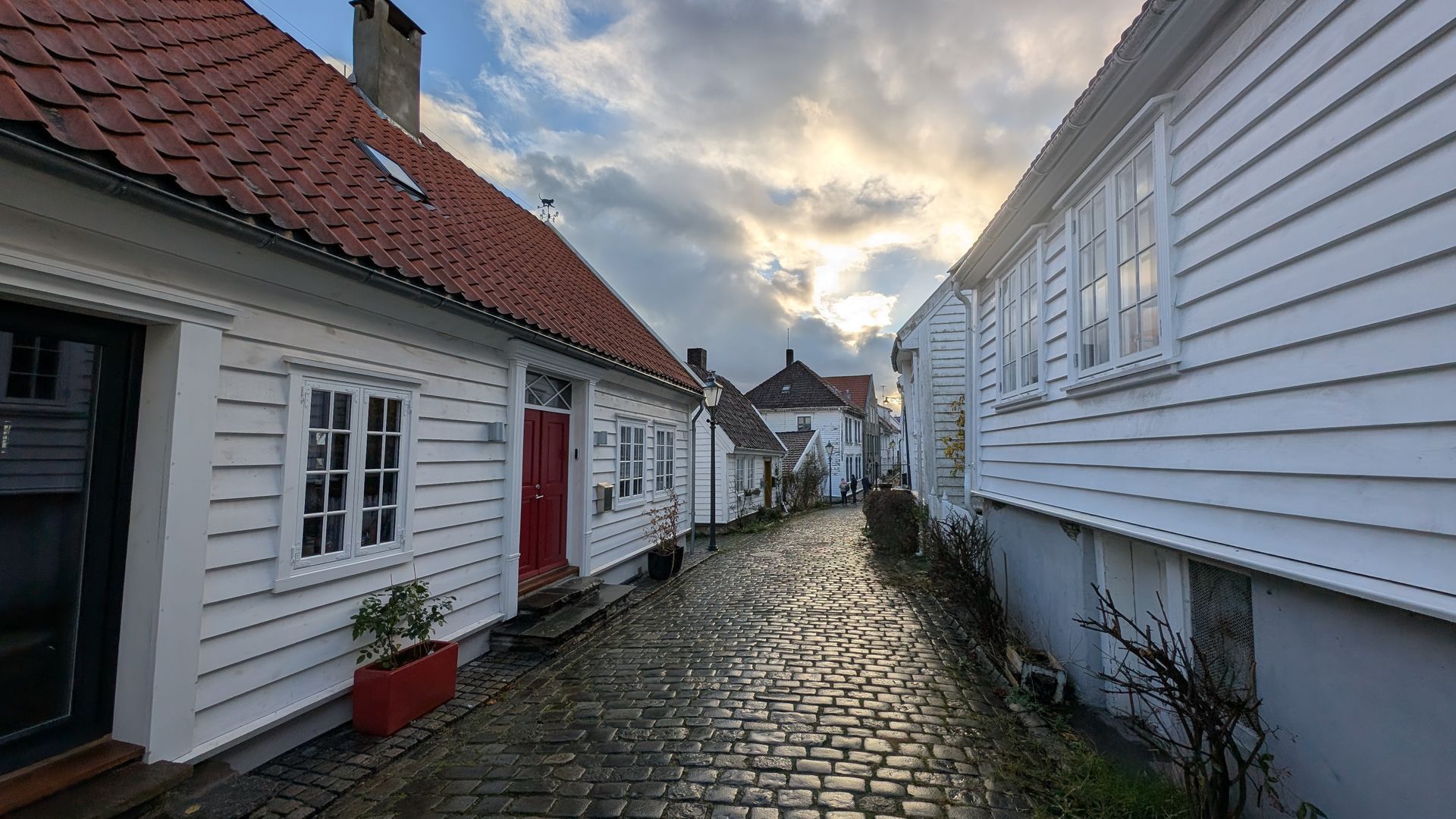 Schmale Kopfsteinpflasterstraße zwischen weißen Holzhäusern mit roten Ziegeldächern. Bewölkter Himmel.