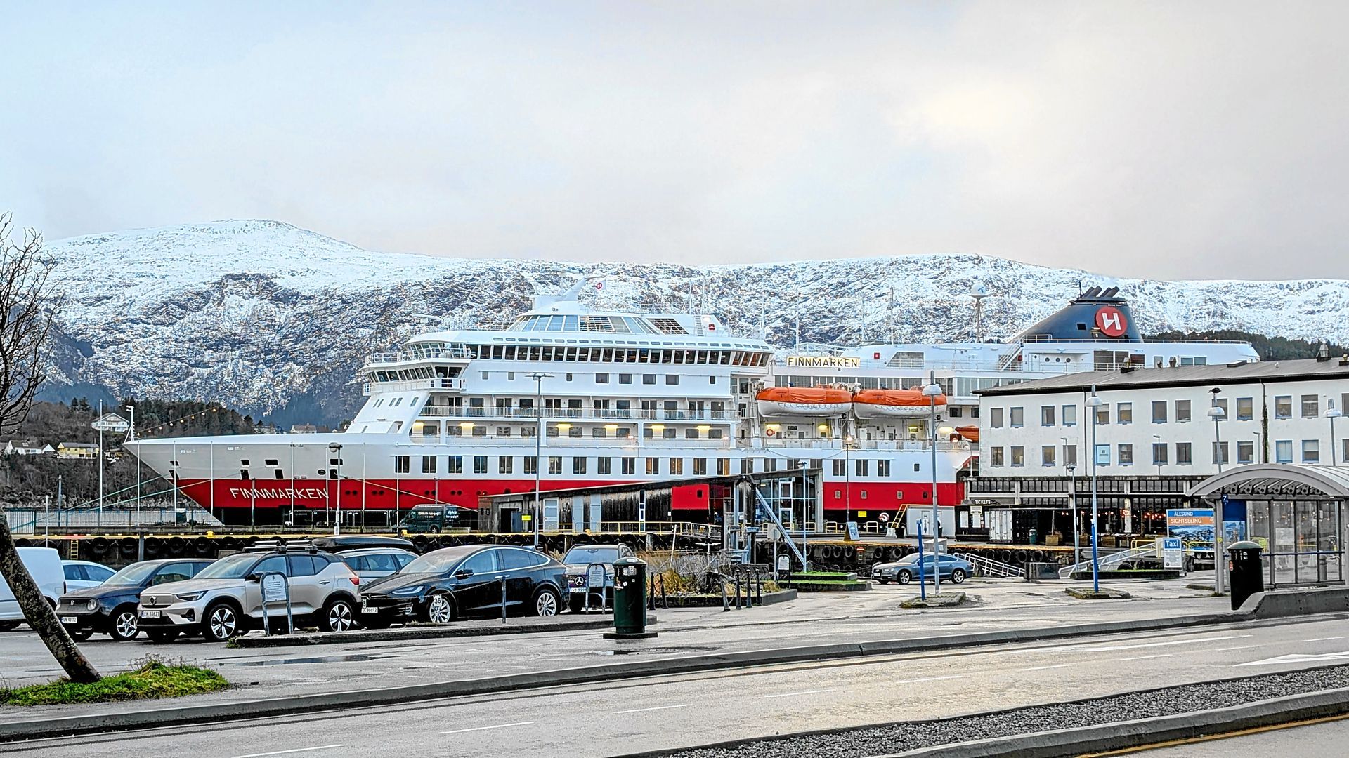 Ein Kreuzfahrtschiff liegt im Hafen vor Anker, rot und weiß vor einer Bergkulisse. Davor parken Autos, im Hintergrund schneebedeckte Berge.