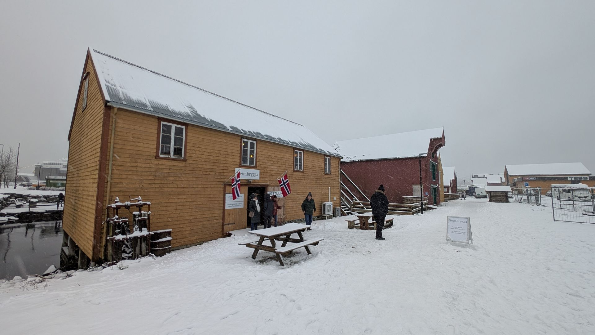 Verschneite Szene: Holzgebäude mit Menschen davor, britische Flagge, Picknicktische.