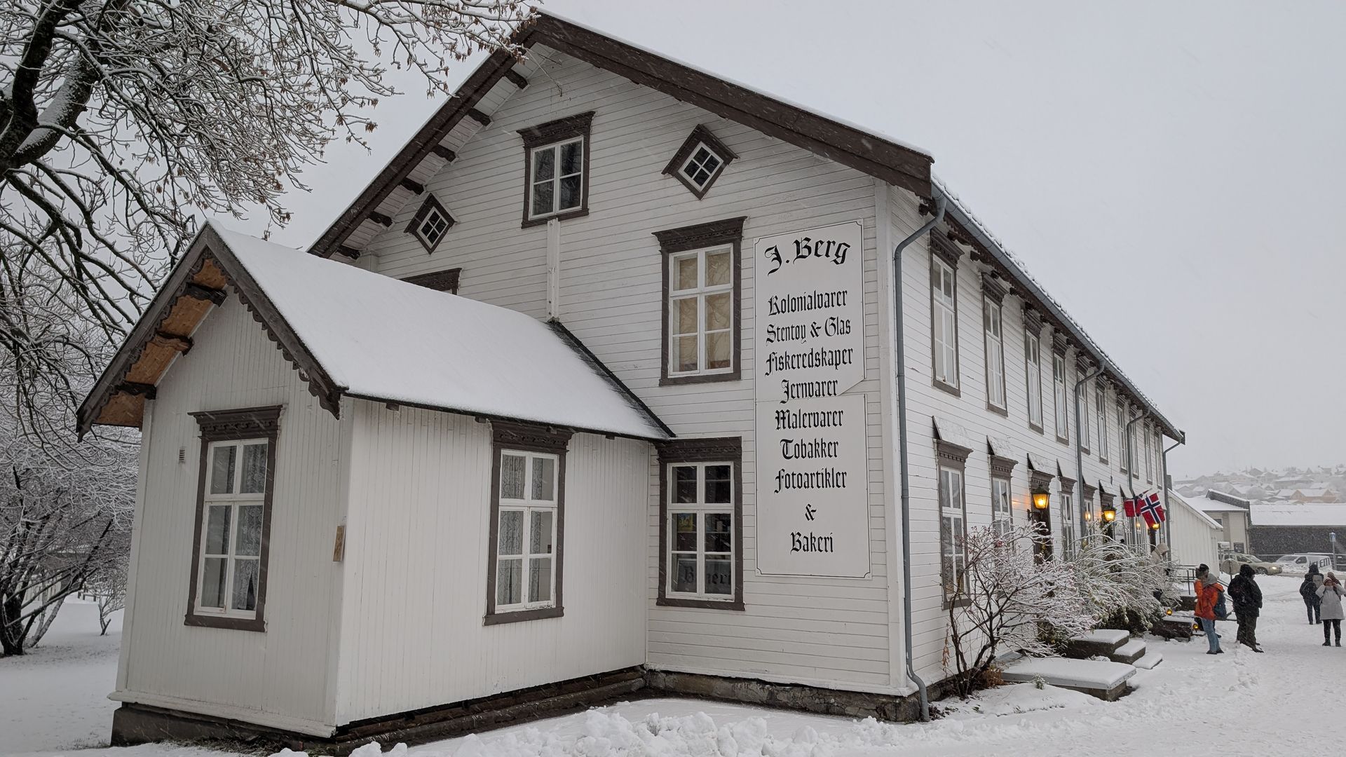 Weißes, zweistöckiges Gebäude im Schnee. Text an der Seite. Menschen in der Nähe der Treppe. Verschneite Bäume und Boden.