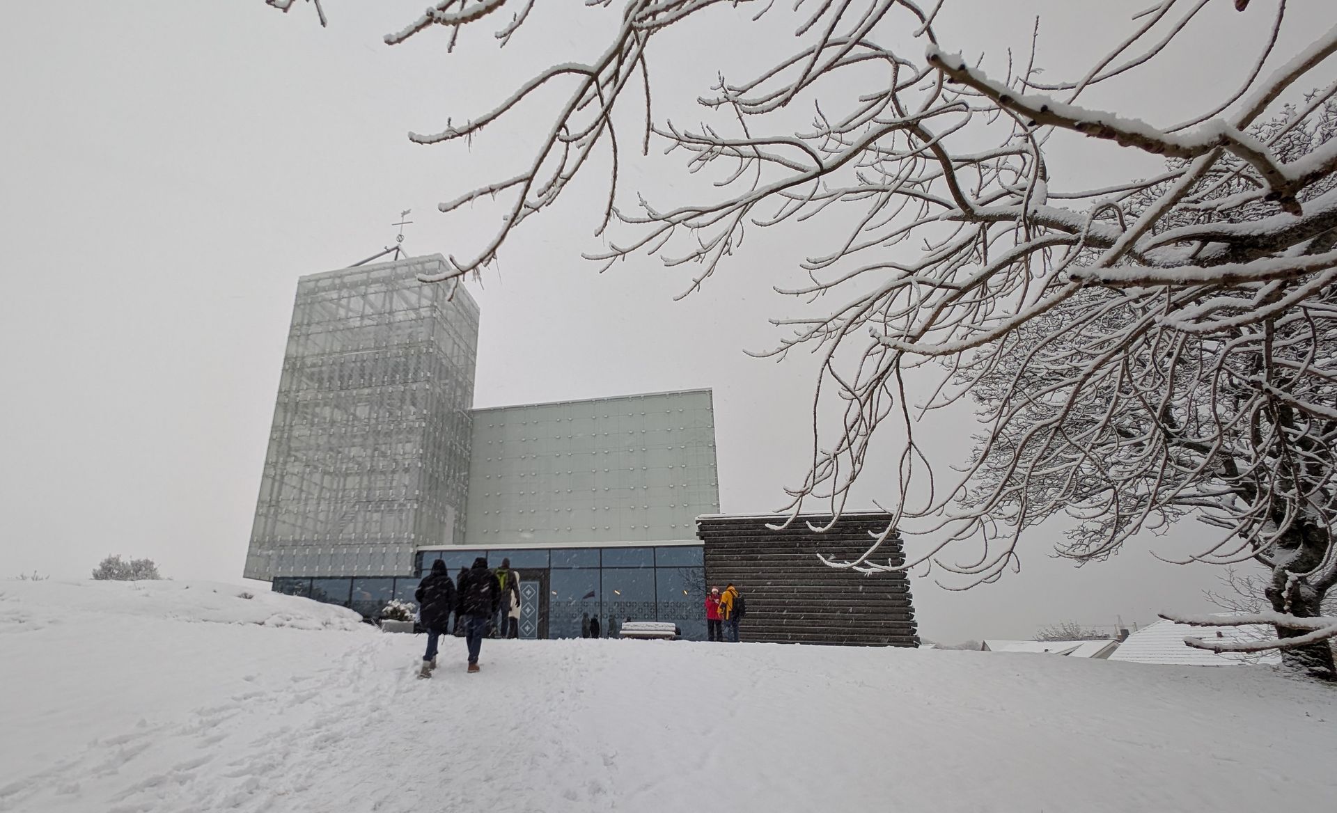 Verschneite Fassade eines modernen Gebäudes mit Turm; Menschen gehen auf den Eingang zu. Im Vordergrund kahle Baumzweige.