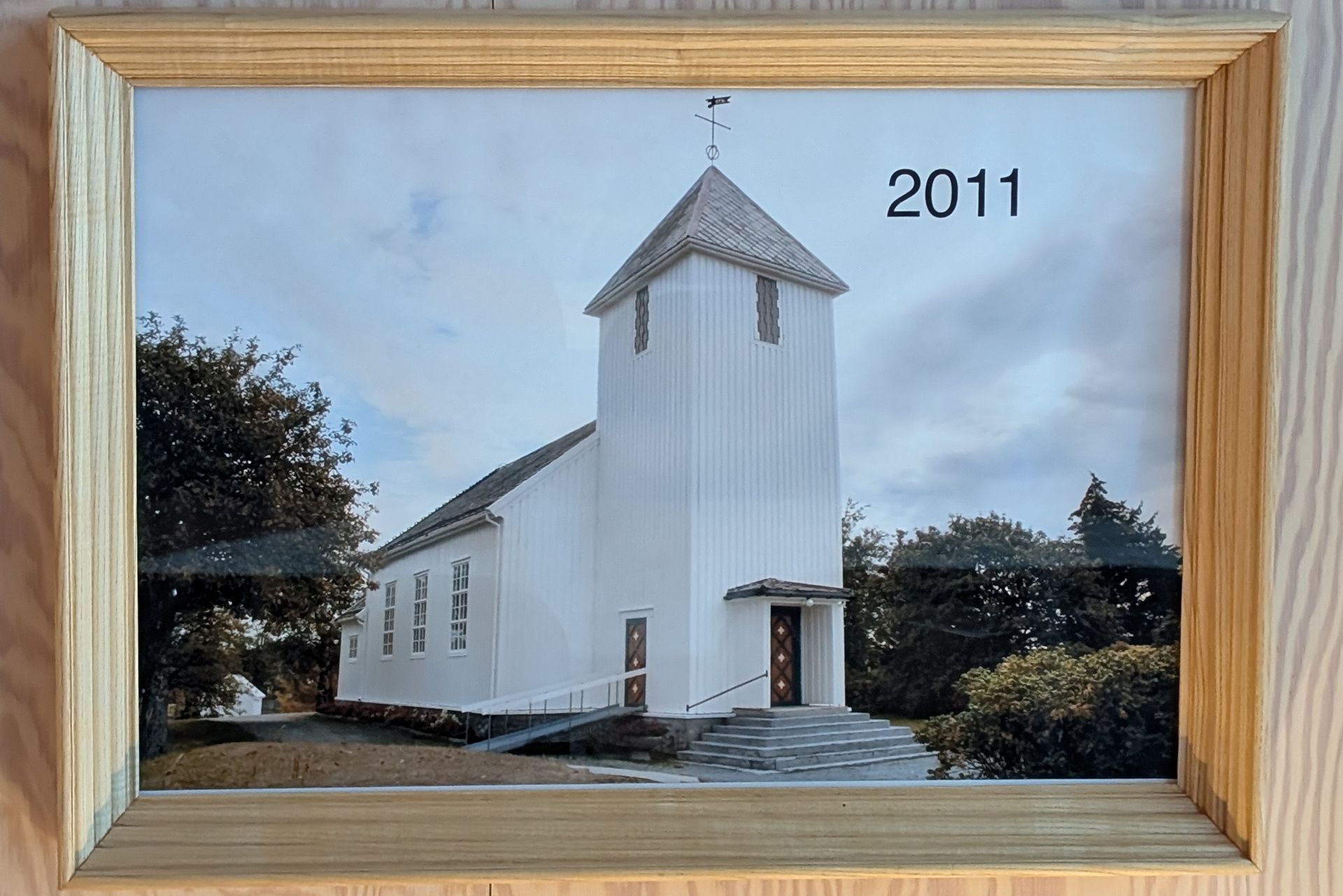 Weiße Kirche in Holzrahmenbauweise, aufgenommen im Jahr 2011. Turm, Bäume und grauer Himmel sind zu sehen.