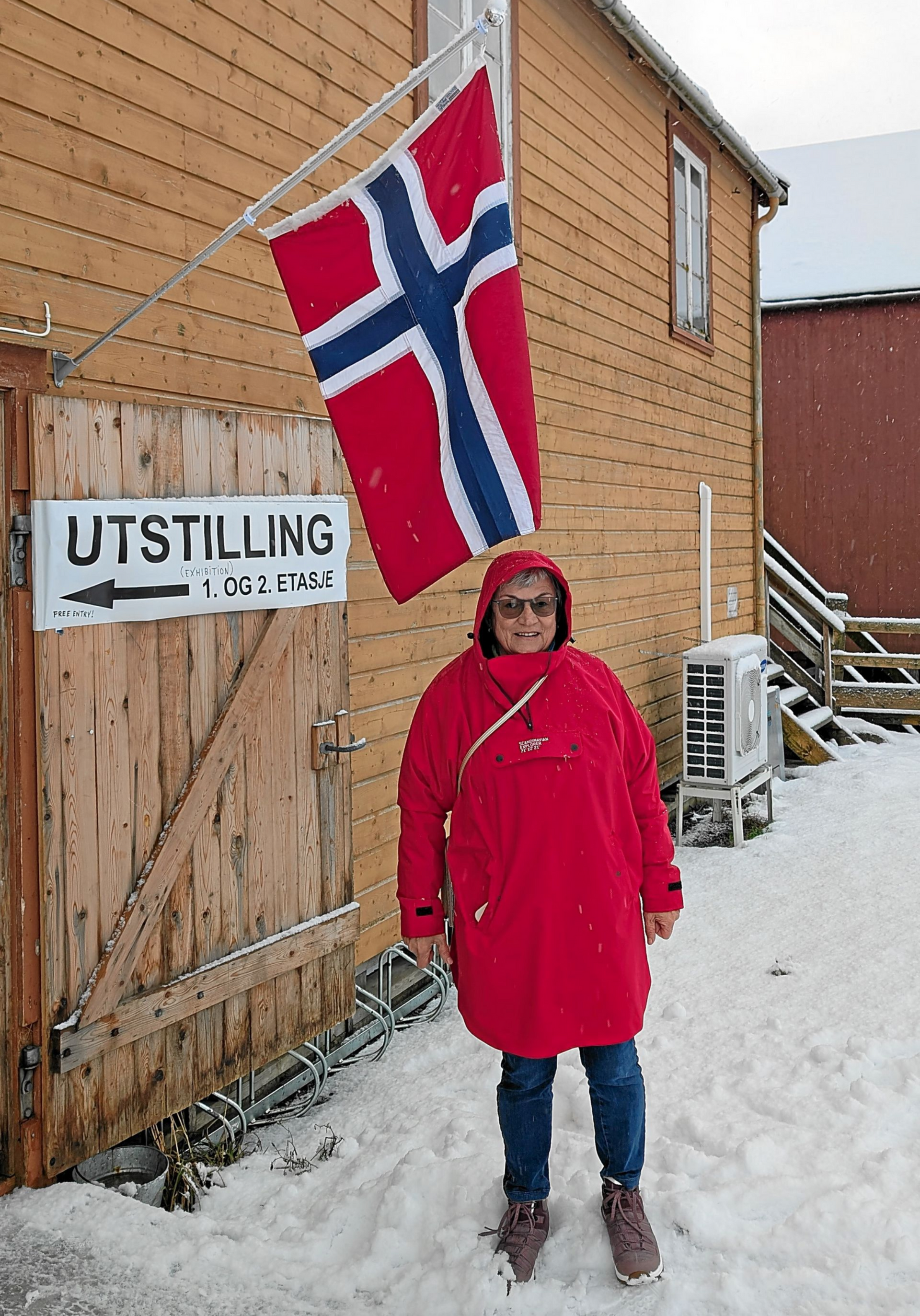 Eine Frau in rotem Mantel steht im Schnee neben einem Holzgebäude mit norwegischer Flagge.
