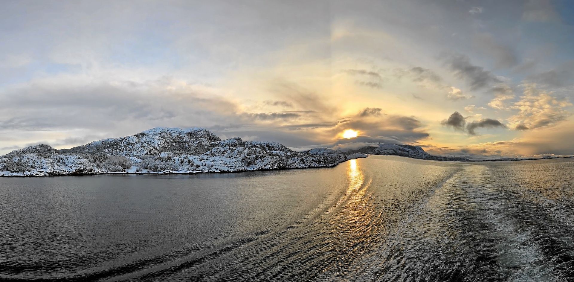 Schneebedeckte Berge und Wasser unter einem bewölkten Himmel, auf dessen Wasseroberfläche sich das Sonnenlicht spiegelt.