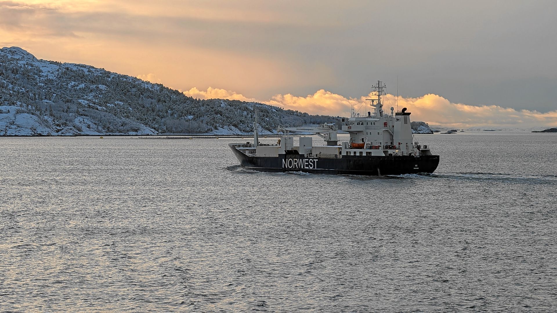 Fähre auf ruhigem Wasser mit schneebedeckten Bergen im Hintergrund unter bewölktem Himmel.