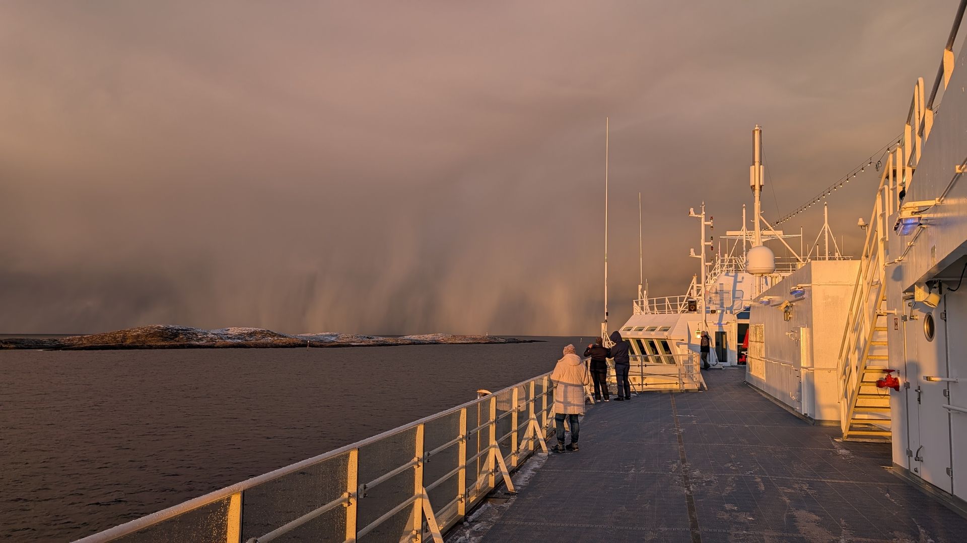 Ein Schiffsdeck, auf dem Menschen bei Sonnenuntergang einen fernen Regenbogen beobachten.