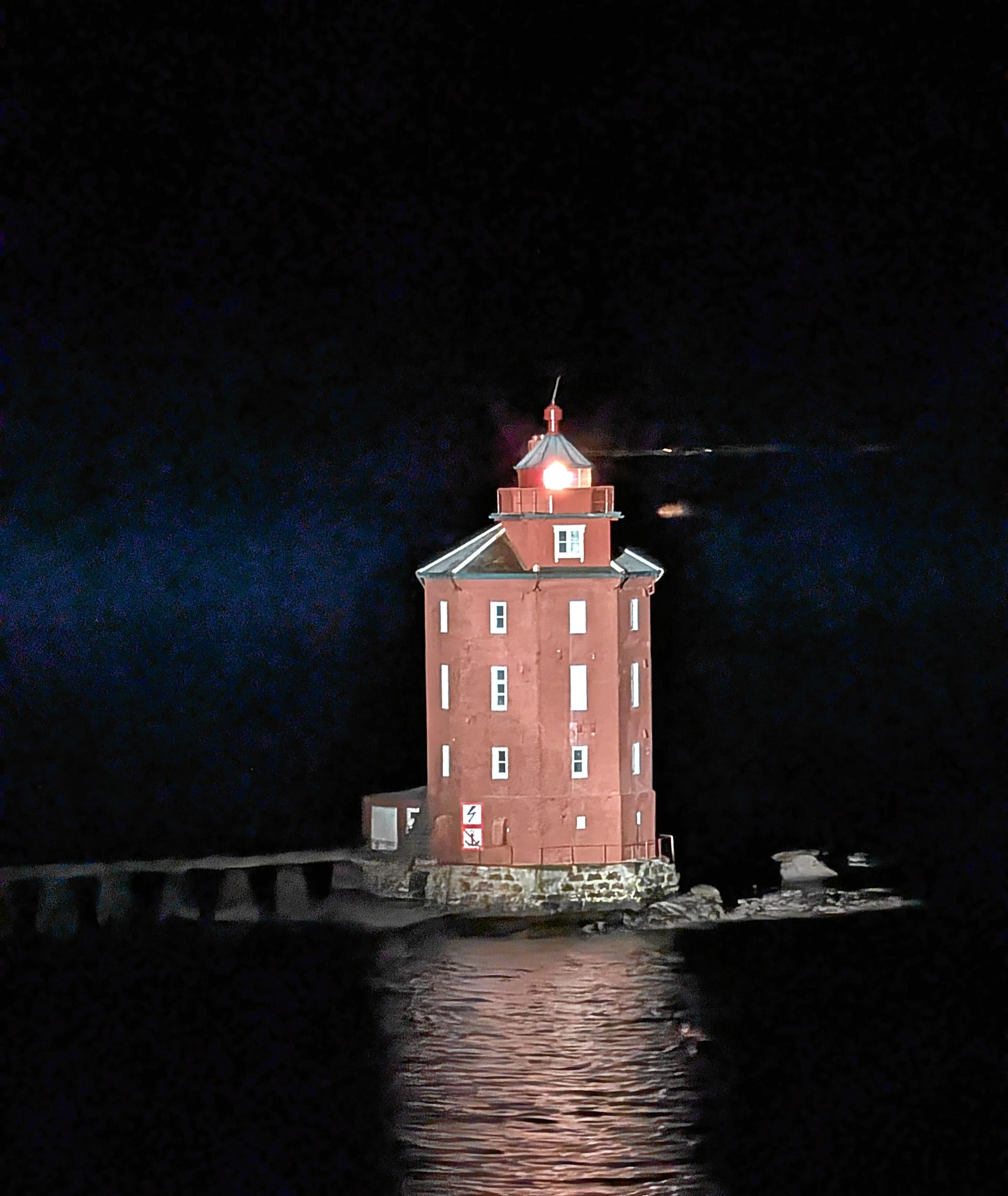 Ein Leuchtturm aus roten Backsteinen mit leuchtender Lampe spiegelt sich in der ruhigen Wasseroberfläche einer dunklen Nacht.
