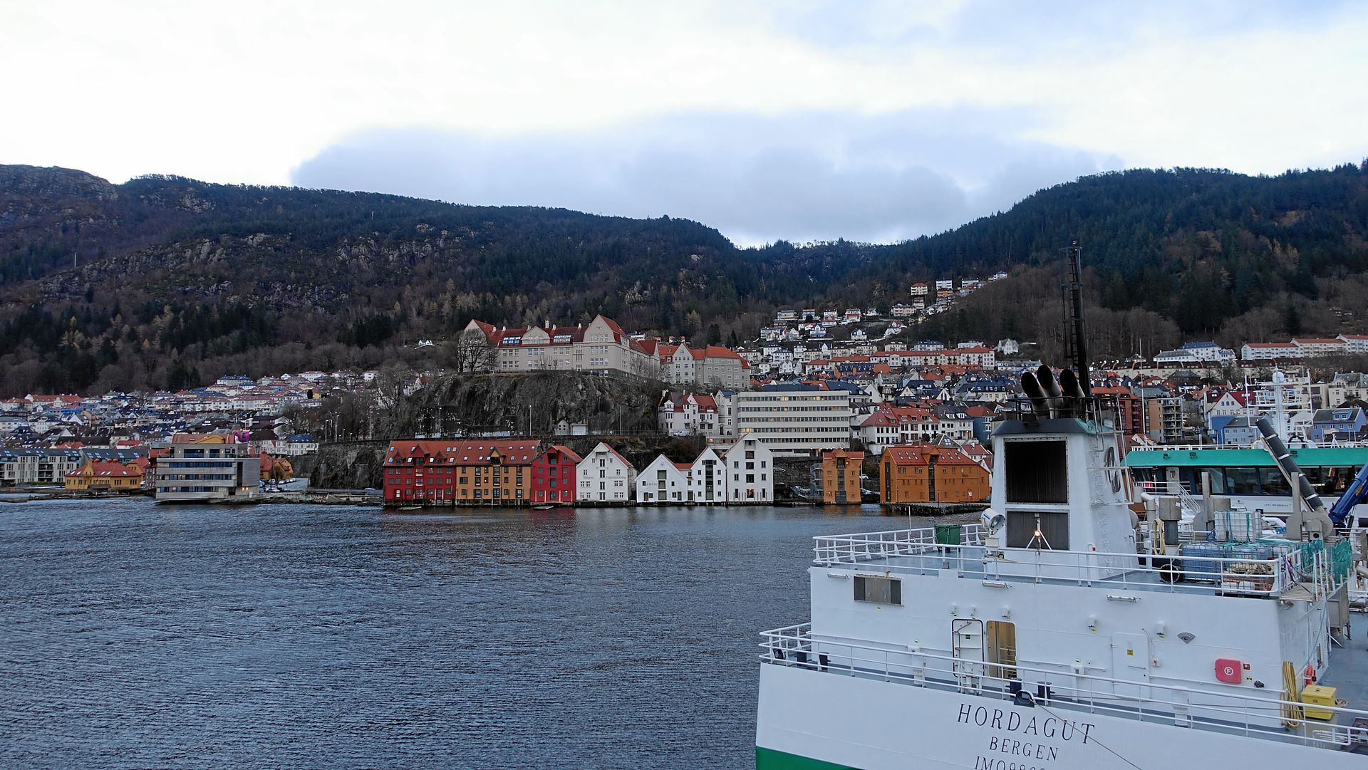 Hafenblick auf Bergen, Norwegen, mit farbenfrohen Gebäuden entlang der Uferpromenade und bewaldeten Hügeln im Hintergrund.