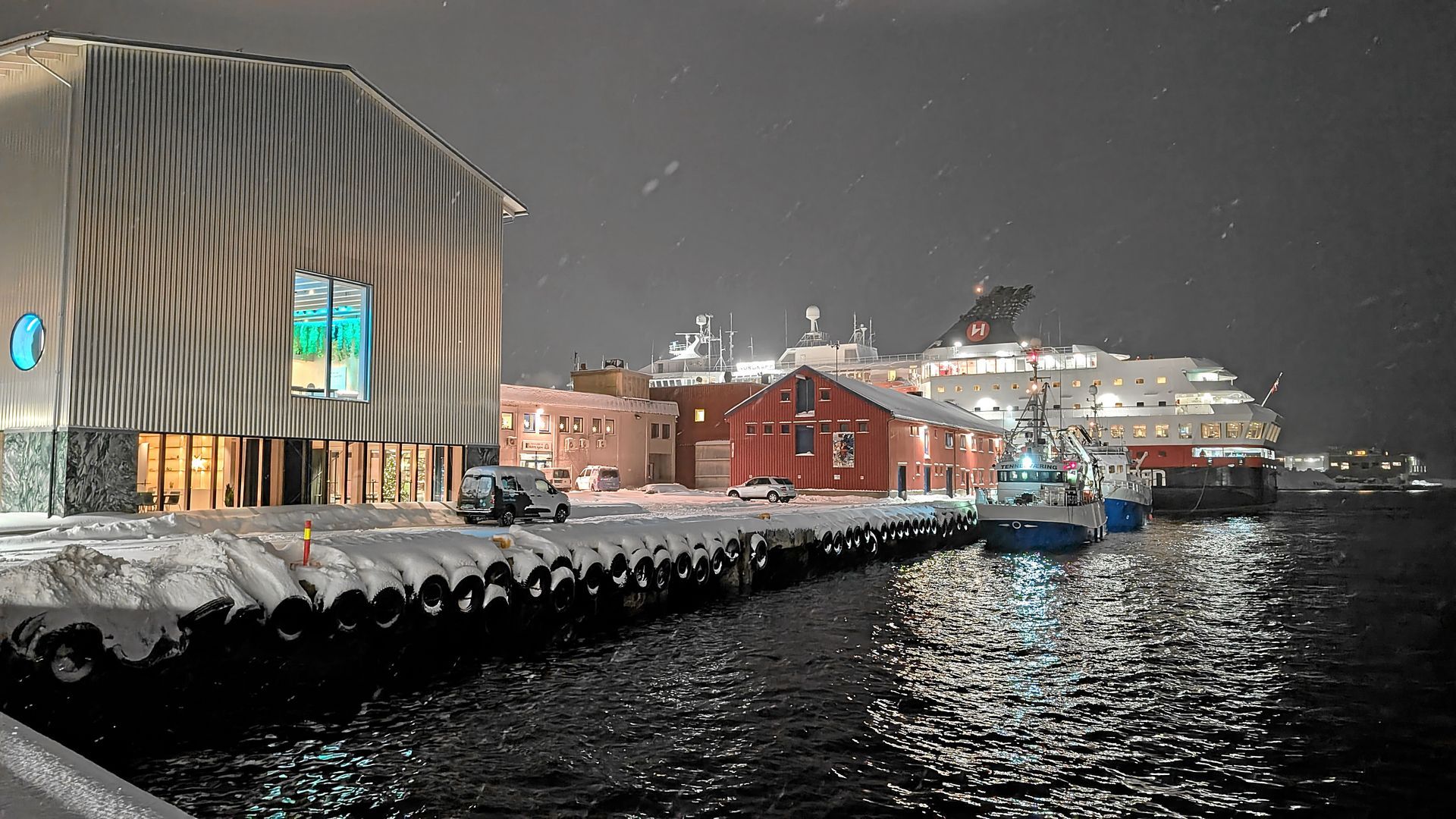 Verschneiter Hafen bei Nacht; ein großes Kreuzfahrtschiff liegt vor Anker, davor ein Gebäude und daneben ein kleines Boot.