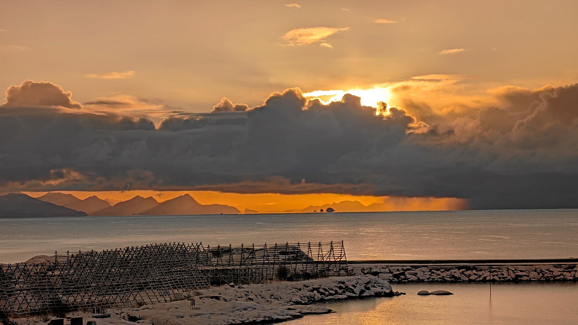 Goldener Sonnenuntergang über dem Wasser und fernen Bergen, mit Wolken und einer Küstenlinie im Blickfeld.