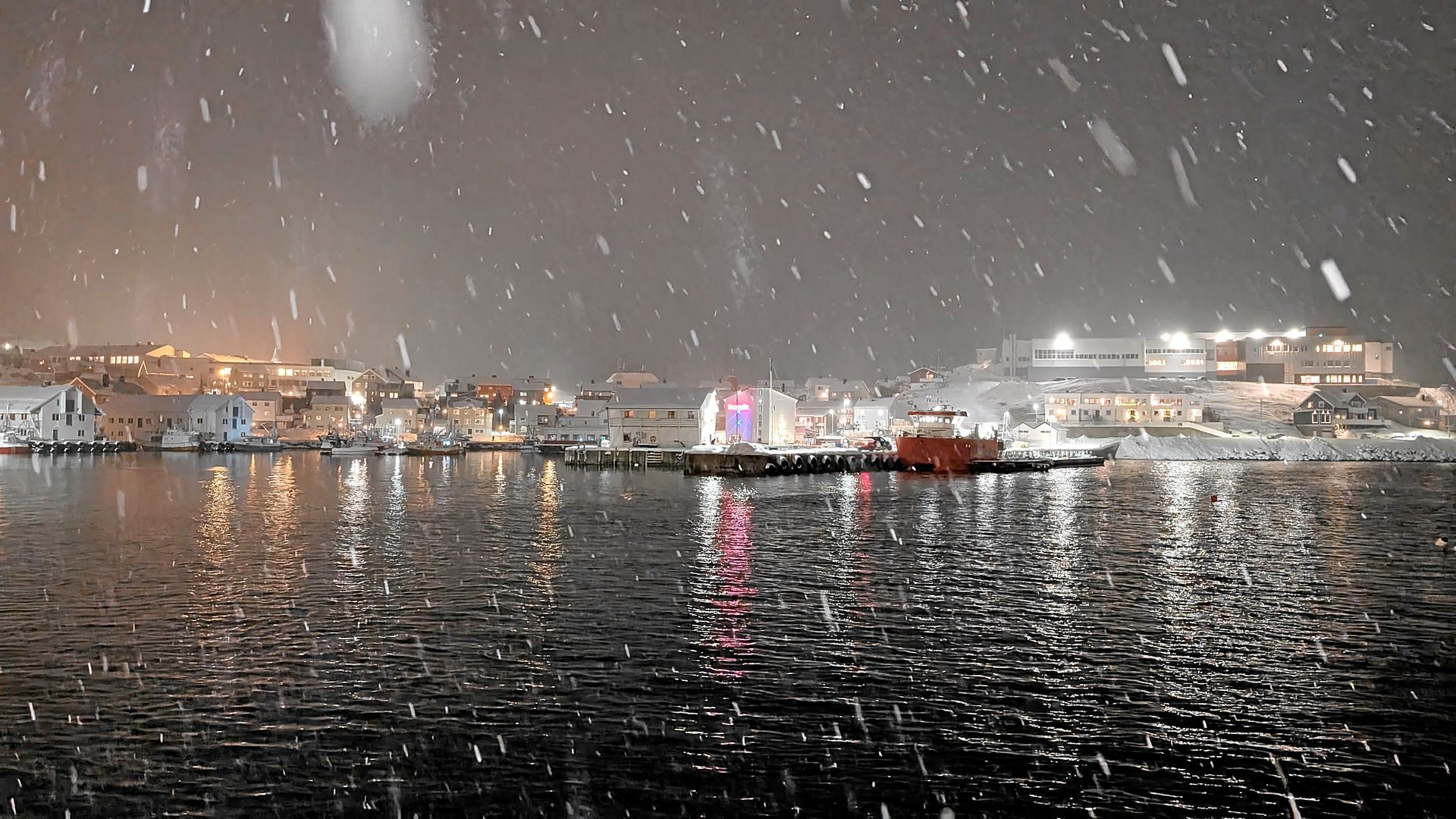 Schnee fällt über einem Hafen, in dessen Wasser sich die Lichter der Stadt spiegeln.