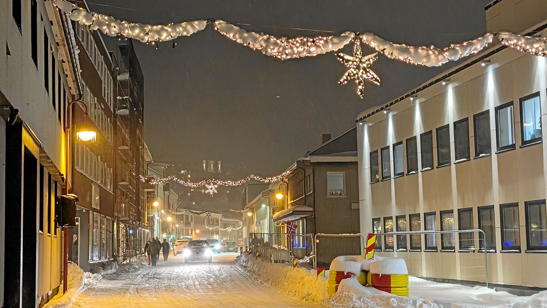 Verschneite Straße mit weihnachtlich beleuchteten Straßenrändern. Gebäude säumen die Straße; einige Fußgänger gehen spazieren.