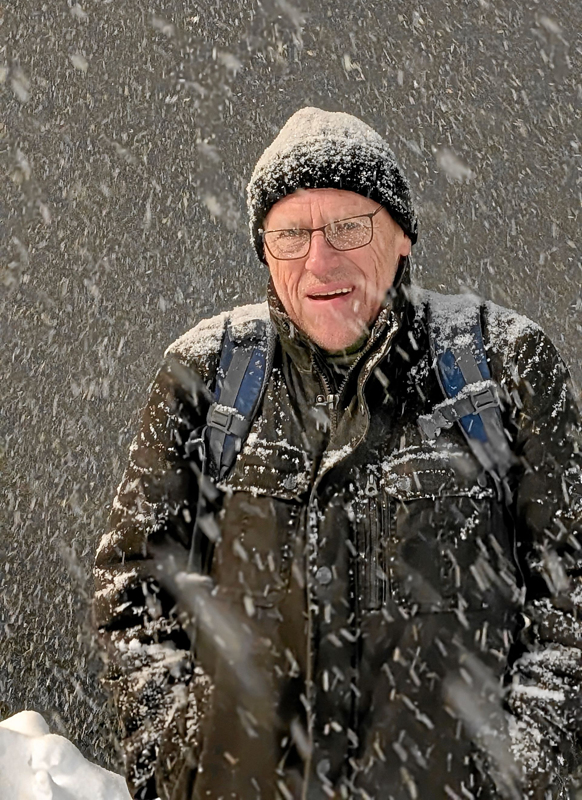 Mann mit Wintermütze und Mantel, in einem Schneesturm, steht in der Nähe einer dunklen Mauer.