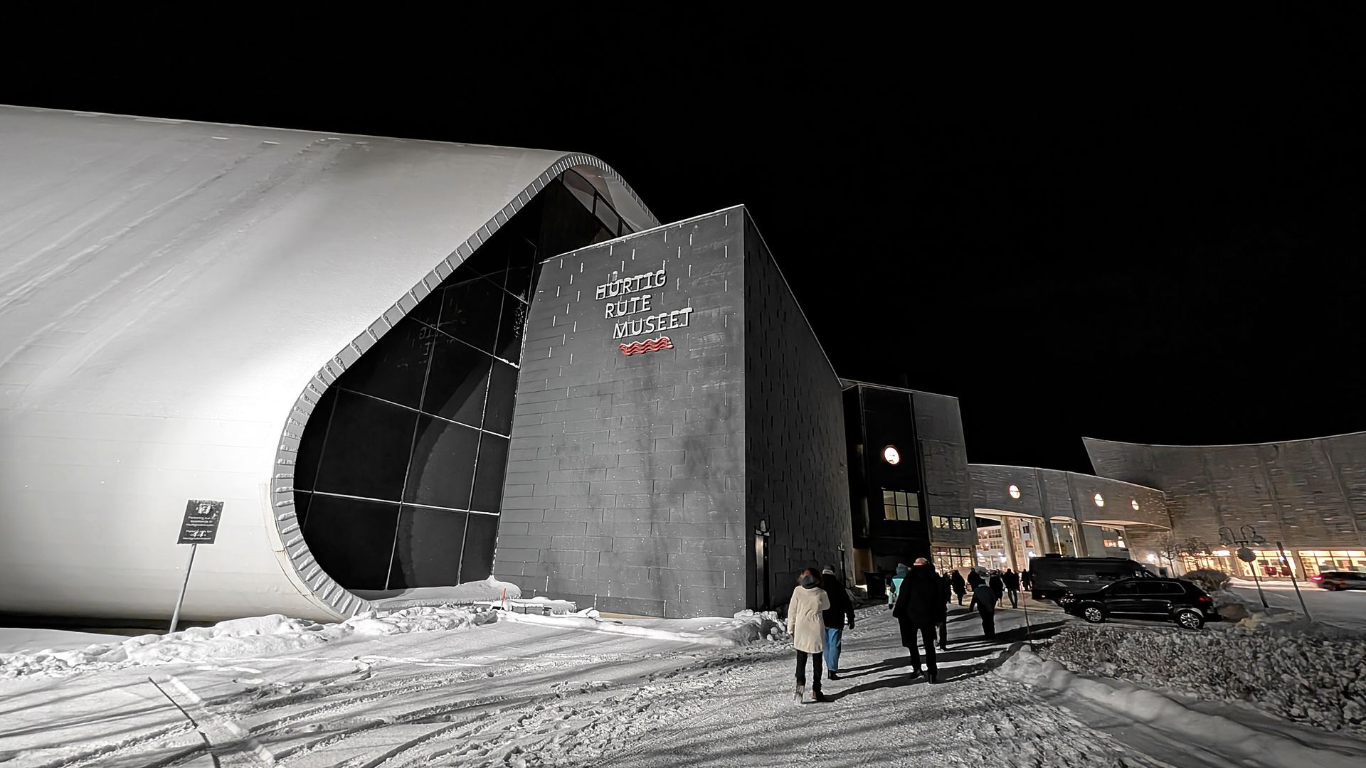 Eingang zu einem modernen Gebäude bei Nacht, Menschen gehen vorbei; schneebedeckter Boden.
