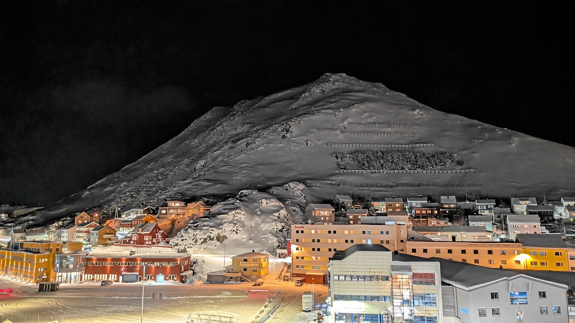 Nachtansicht einer Stadt, die sich an einen schneebedeckten Berg schmiegt, wobei die Lichter der Gebäude die Szene erhellen.