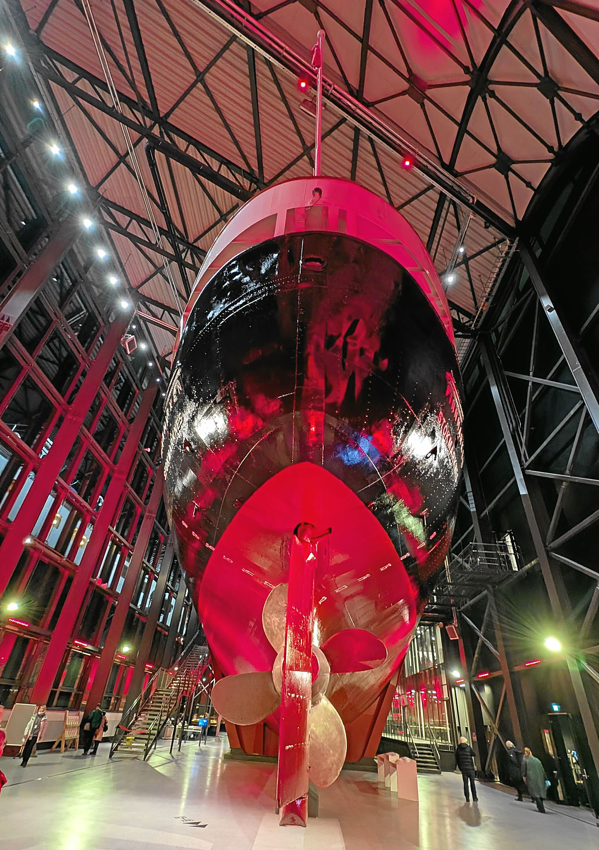 Ein rot beleuchteter Schiffsbug in einer großen Halle, der einen Propeller zeigt und das Licht der Umgebung reflektiert.