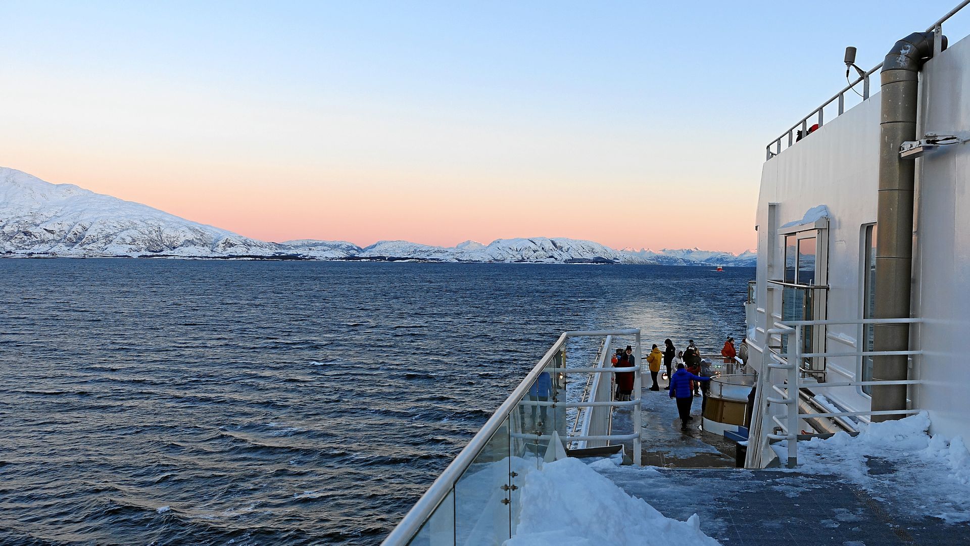 Schiffsdeck mit Schnee, Blick über eisiges Wasser auf schneebedeckte Berge in der Abenddämmerung.
